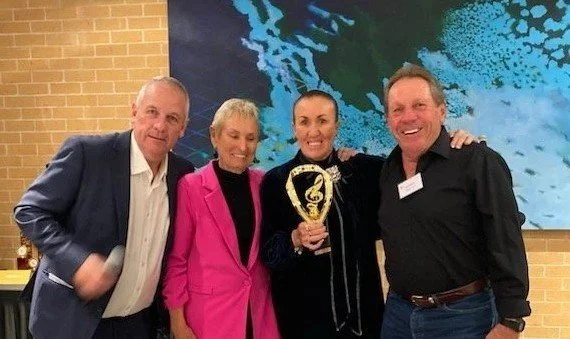 Four people standing together indoors, smiling, with one holding a trophy. Background features a large digital screen with a blue and green abstract design.