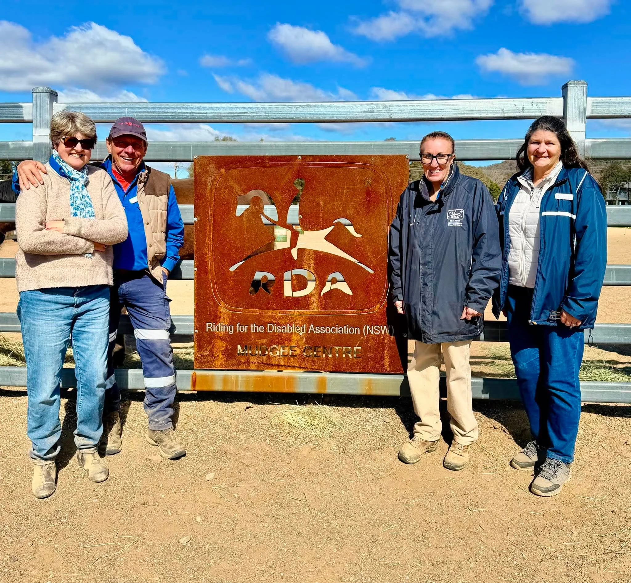 Four women and one man standing outdoors behind a rusted metal sign with the logo and name of the Riding for the Disabled Association (NSW) at Mudgee Centre, smiling at the camera on a sunny day with a blue sky and some clouds.