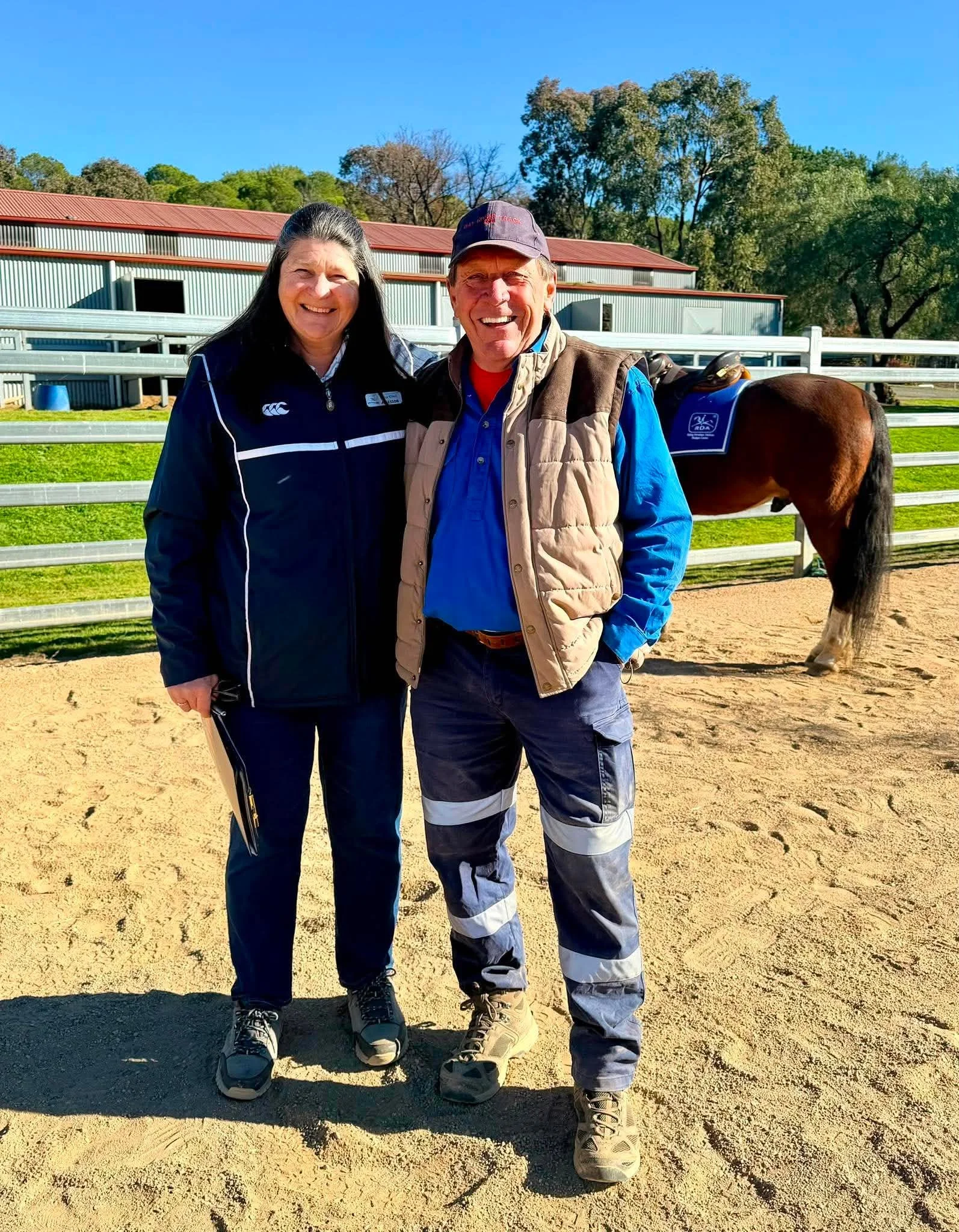 A woman and a man smiling and standing together outdoors on a sunny day, with a horse in the background on a farm or stable.