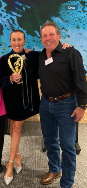 Two smiling people, a woman holding an award, and a man standing next to her with his arm around her, in an indoor setting with a blue tree-themed backdrop.