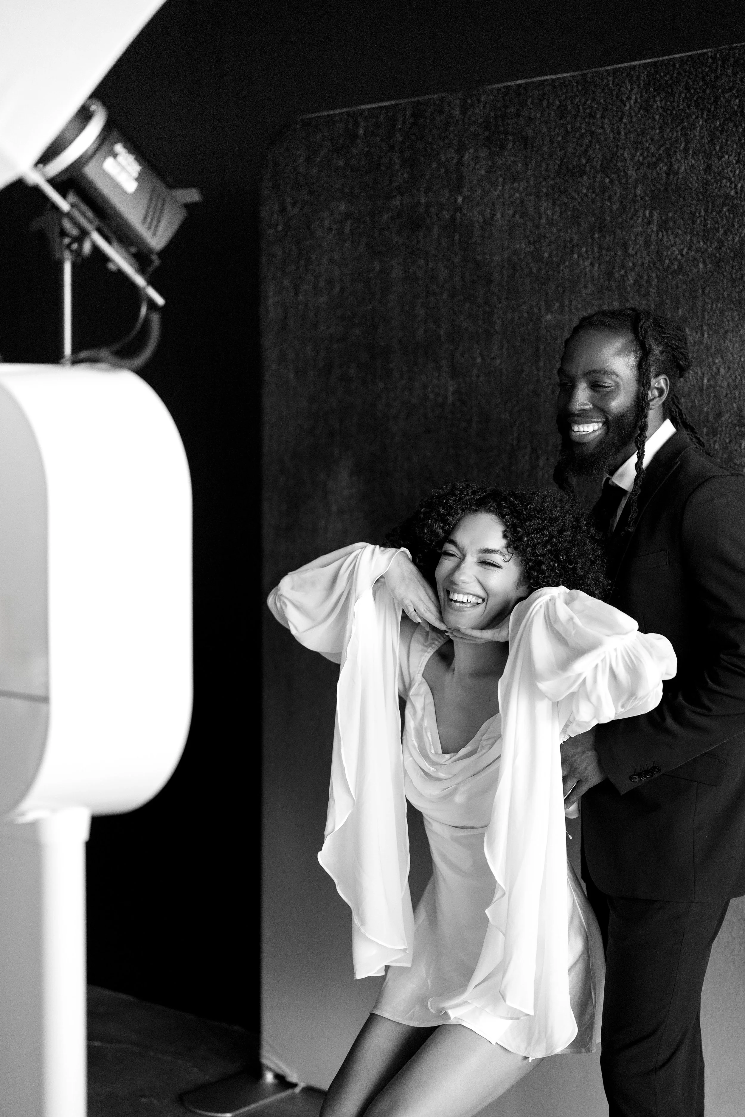 Two people in formal attire laughing and taking a selfie in a photo studio