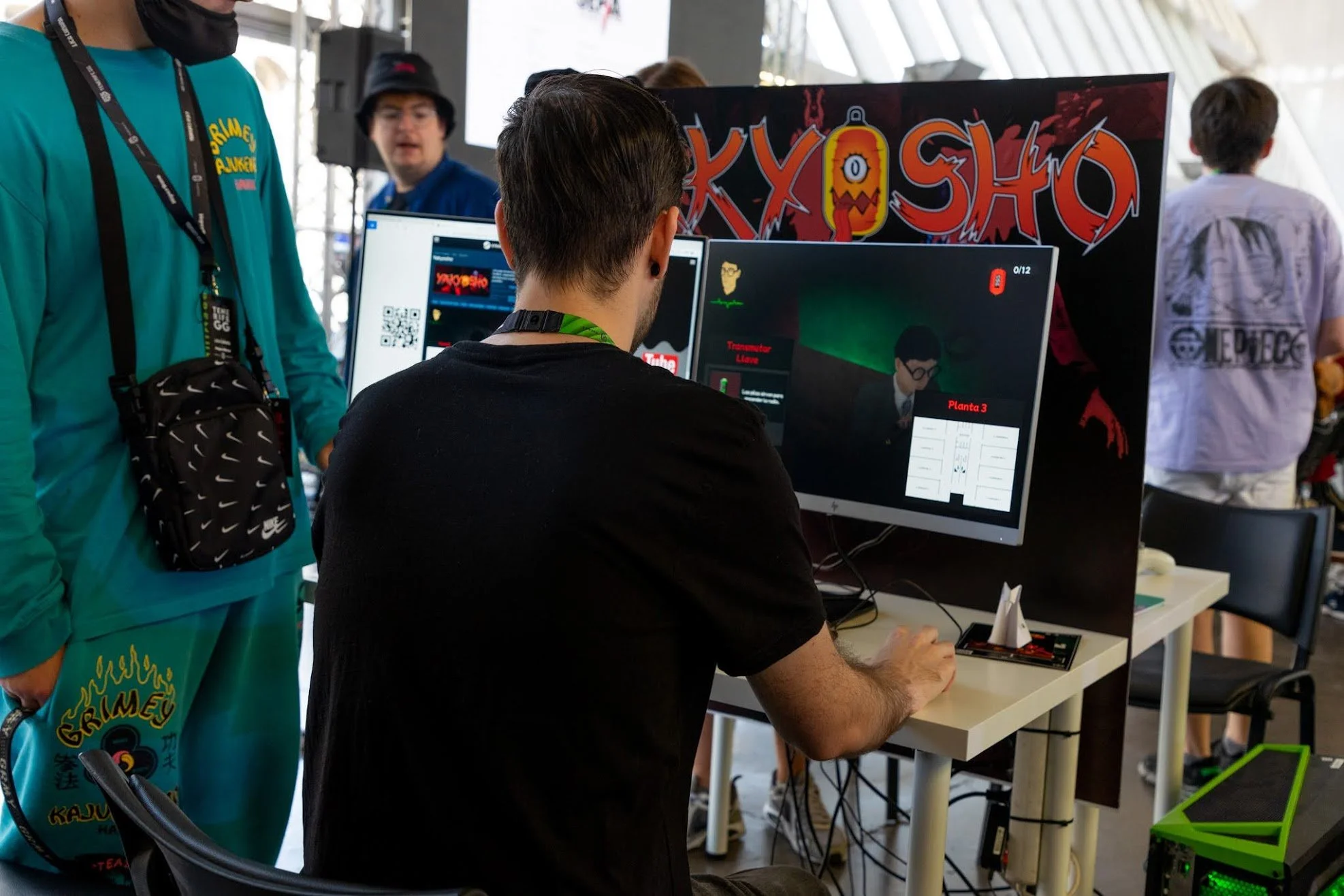 A person with dark hair and black t-shirt working on a computer with dual monitors, with a colorful "KYO SHO" banner in the background at a gaming or technology event.