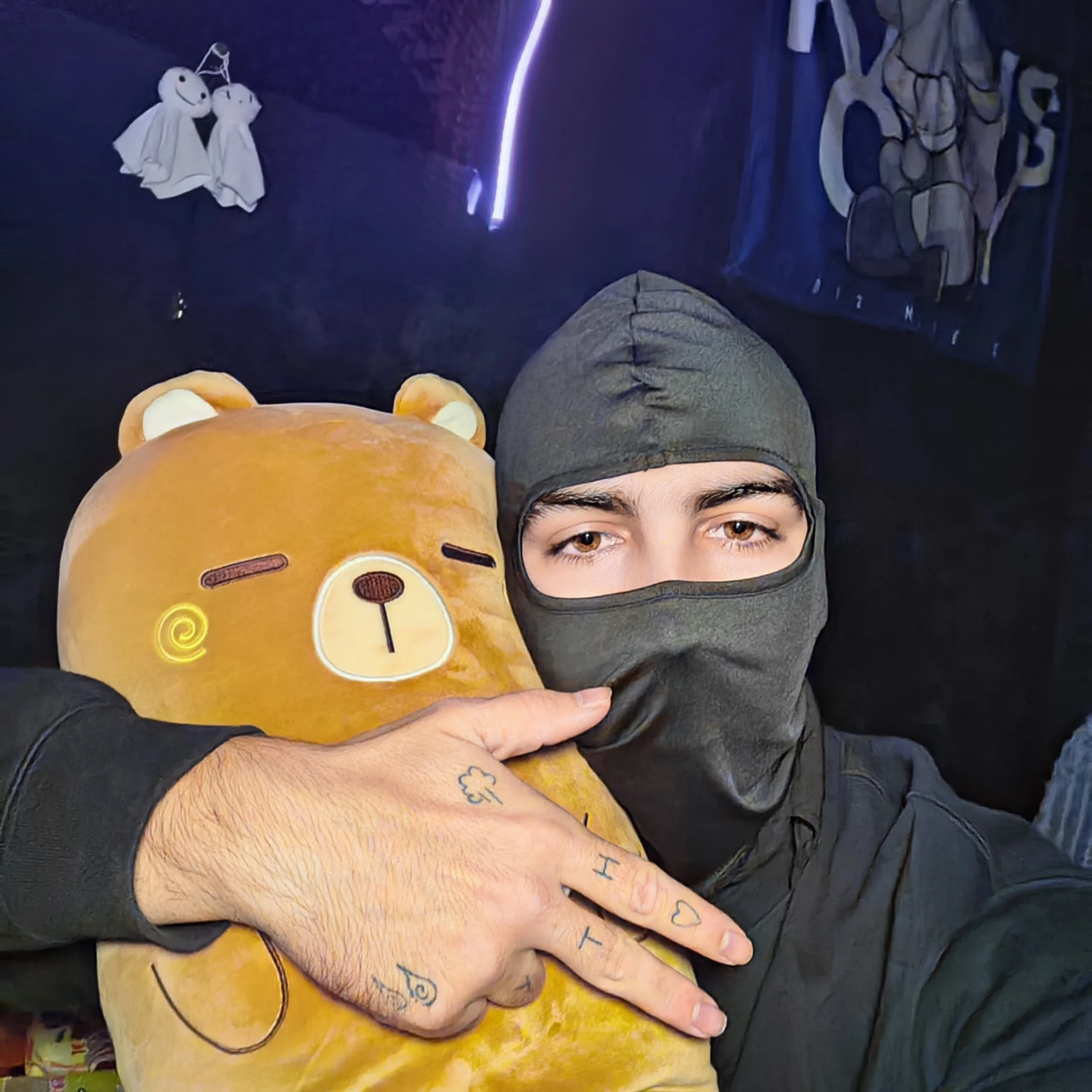 A person wearing a black face mask and a dark hoodie holds a large, plush bear with a winking face. The person's hand gestures a peace sign, showing tattoos on fingers and knuckles. The background is dark with some hanging fabrics and light streaks.