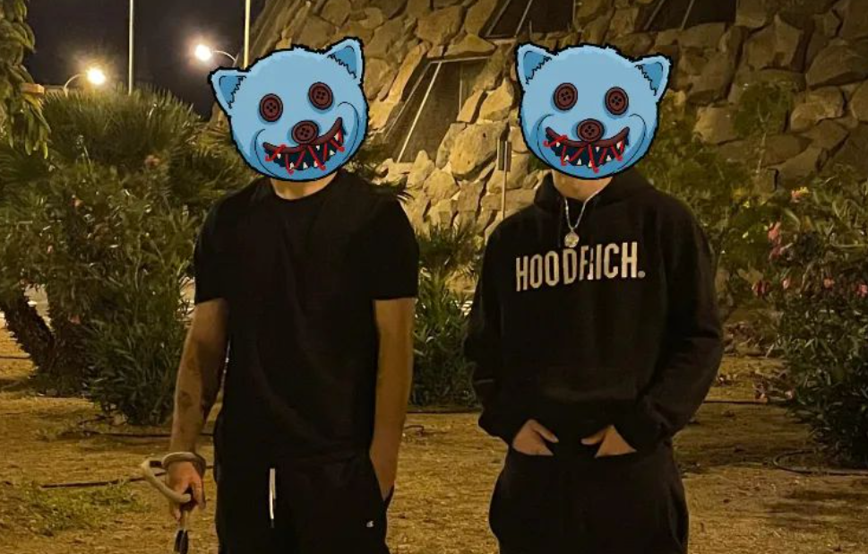 Two people standing outdoors at night with illuminated bushes and a stone wall behind them, their faces obscured by cartoon monster masks.