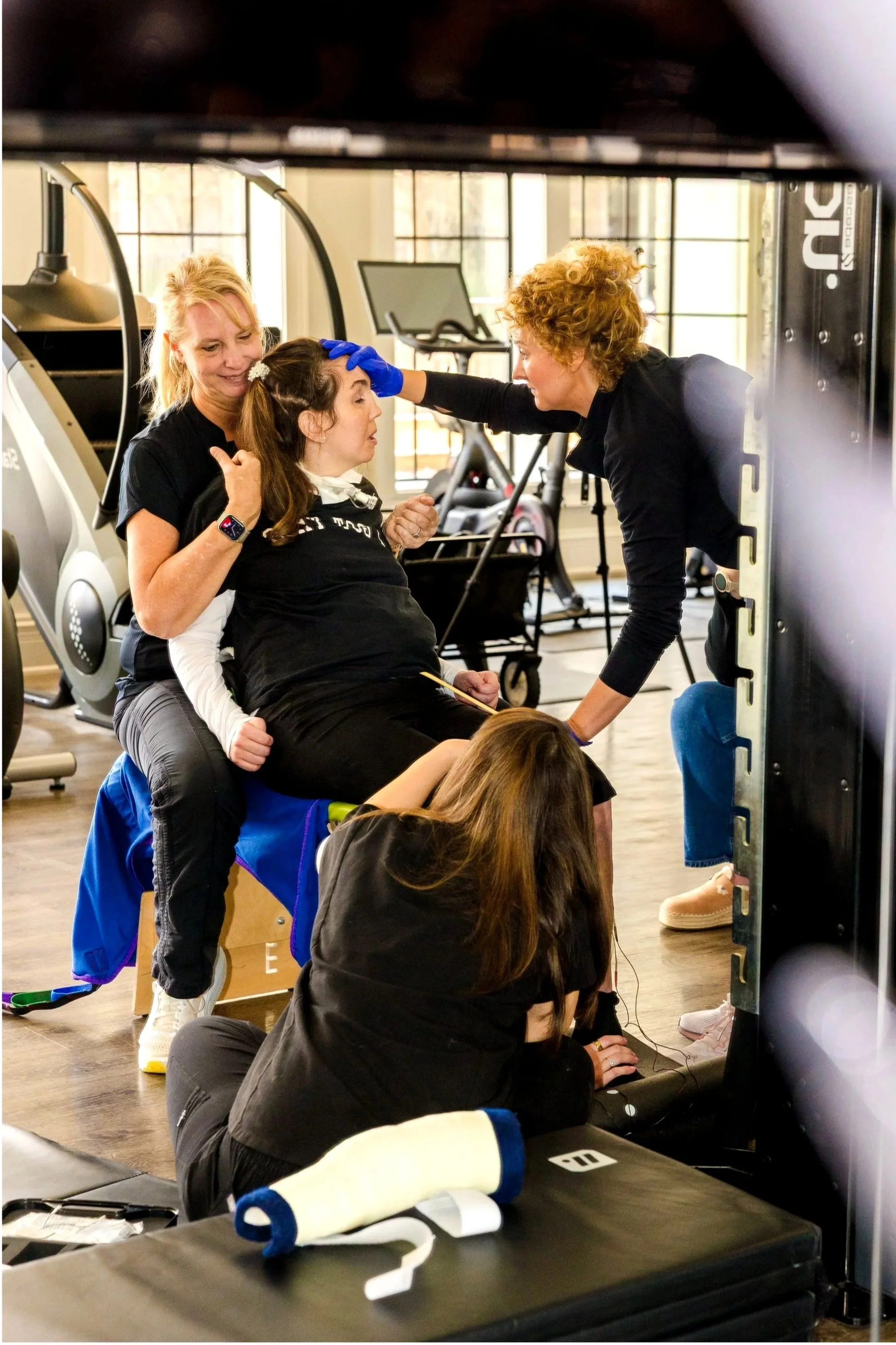 Two women, one with curly blonde hair and one with long brown hair, assist a seated woman with dark hair in a gym or fitness studio. The woman with dark hair is in a recovery position, with her eyes closed and head tilted back as one woman checks her forehead, and the second woman supports her from behind. Several exercise bikes and large windows are in the background.