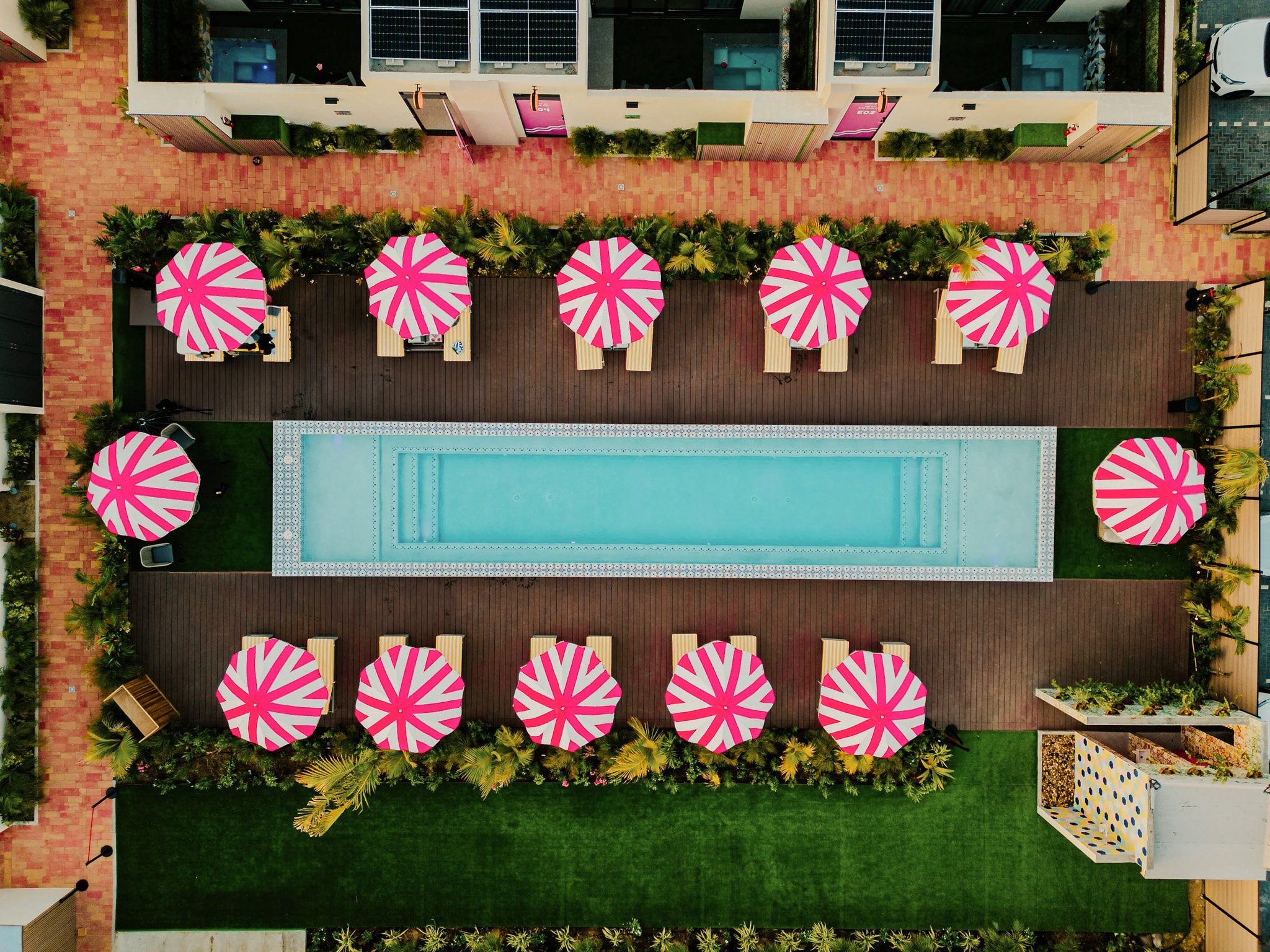 Aerial view of a swimming pool area surrounded by pink and white striped umbrellas, lounge chairs, and green plants.