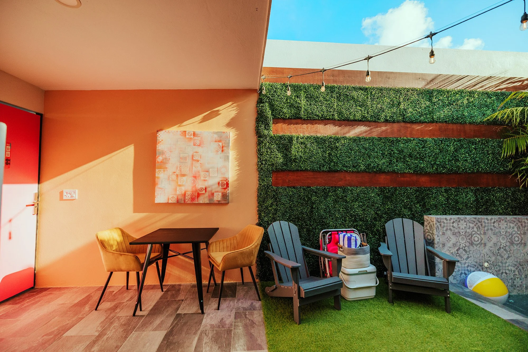 Part of an outdoor patio area with a green wall, string lights, two black Adirondack chairs, and a pool with a yellow and white beach ball.