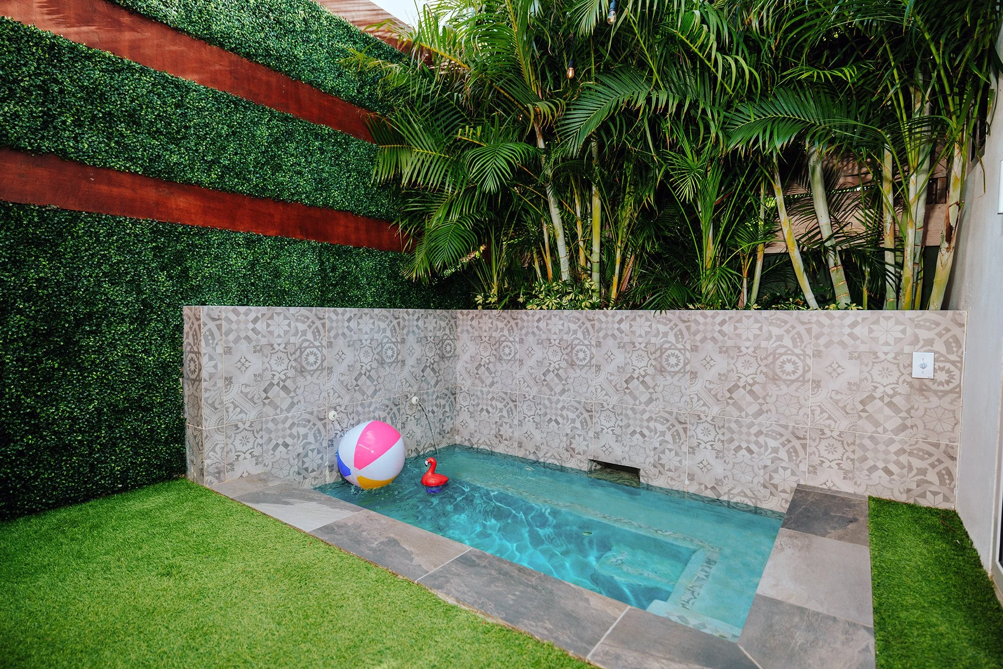 Small backyard pool with a colorful beach ball and a pink flamingo float, surrounded by green grass, a decorative tiled wall, and lush plants.