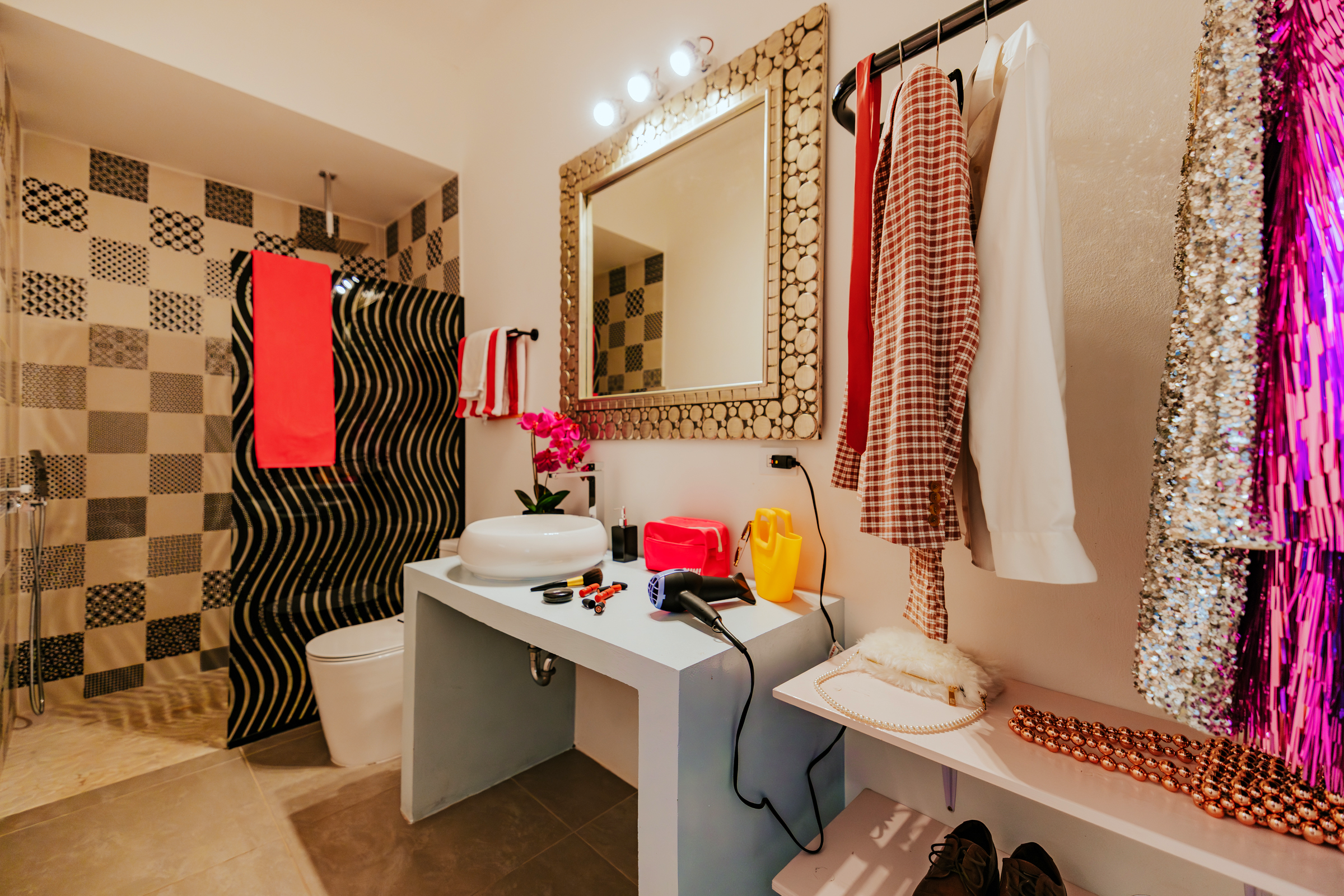 A bathroom with a shower area that has black and beige patterned tiles, a white toilet, a white countertop with a vessel sink, a mirror with a wooden log frame, and various items including a hairdryer, makeup, and clothing hung on a rod.