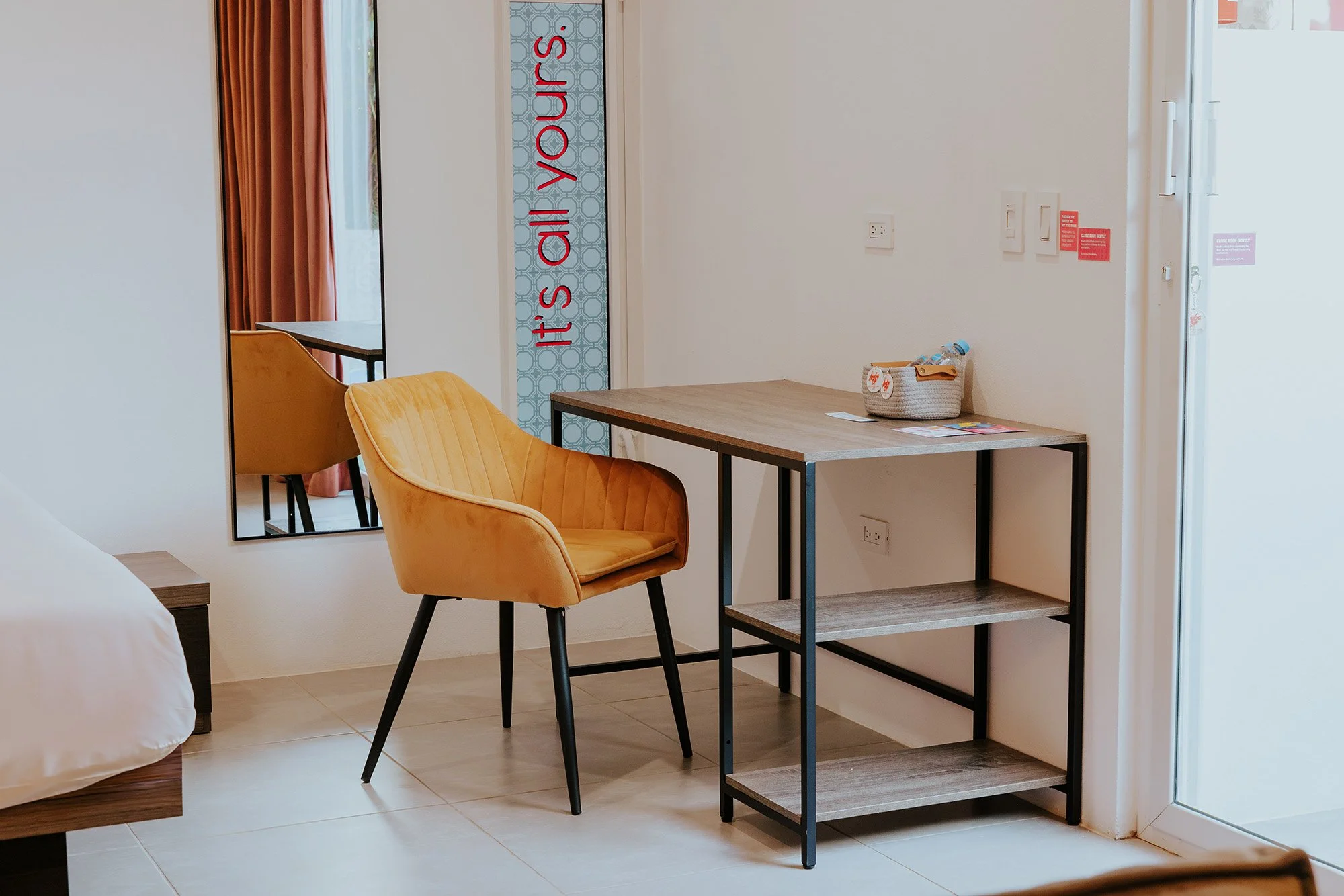 A hotel room with a yellow armchair, a wooden desk with a shelf, a mirror on the wall, and a bed partially visible to the left. The room has white walls, tiled floors, and a glass door leading outside.