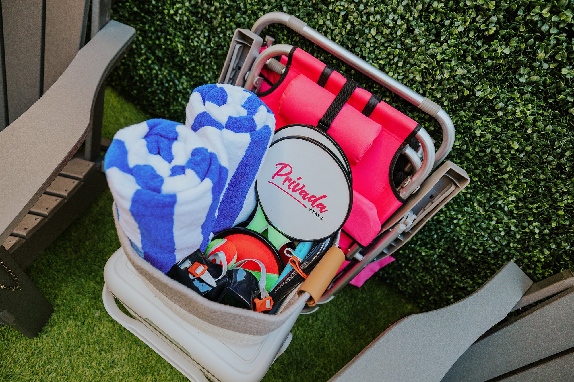 A shopping cart filled with striped blue and white towels, a pink foldable chair, a sign with the word 'Privada' on it, and various small items, situated outdoors on grass next to a bench and a hedge.