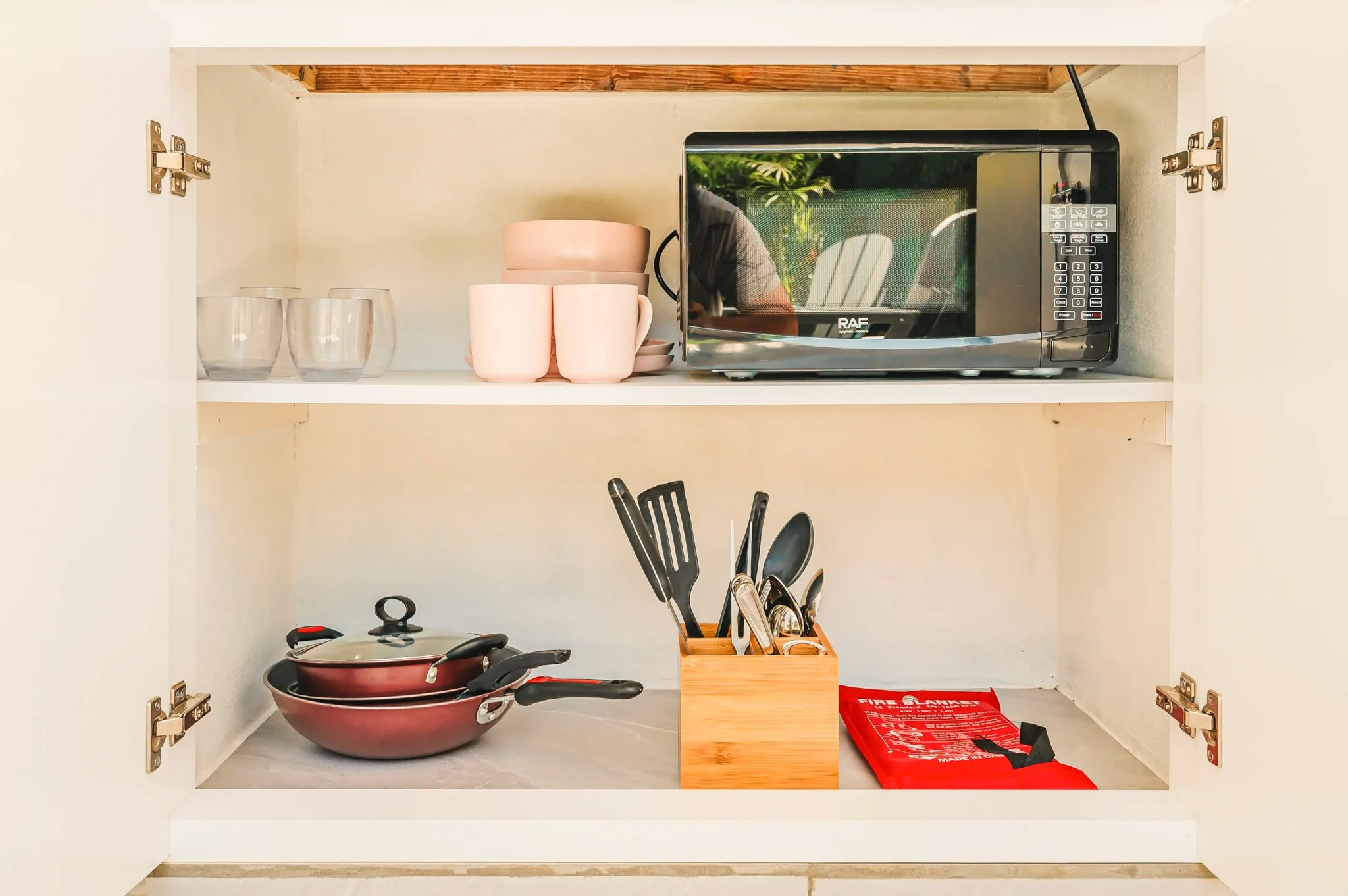 Open kitchen cabinet with glasses, mugs, a microwave, frying pans, utensils, and a fire extinguisher.