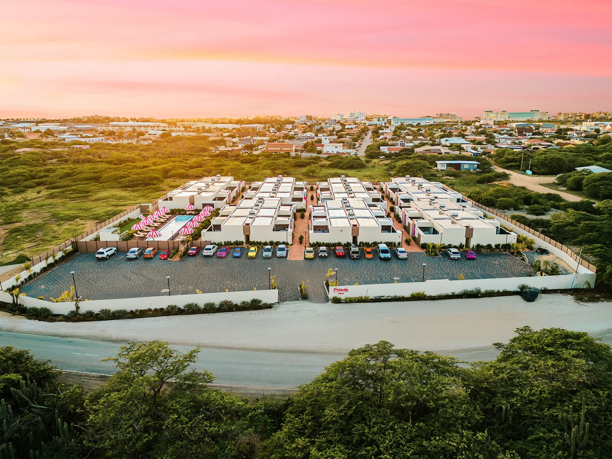 Aerial view of a small residential complex with white units, pink umbrellas, and a parking lot, surrounded by greenery, with a cityscape in the background during sunset.