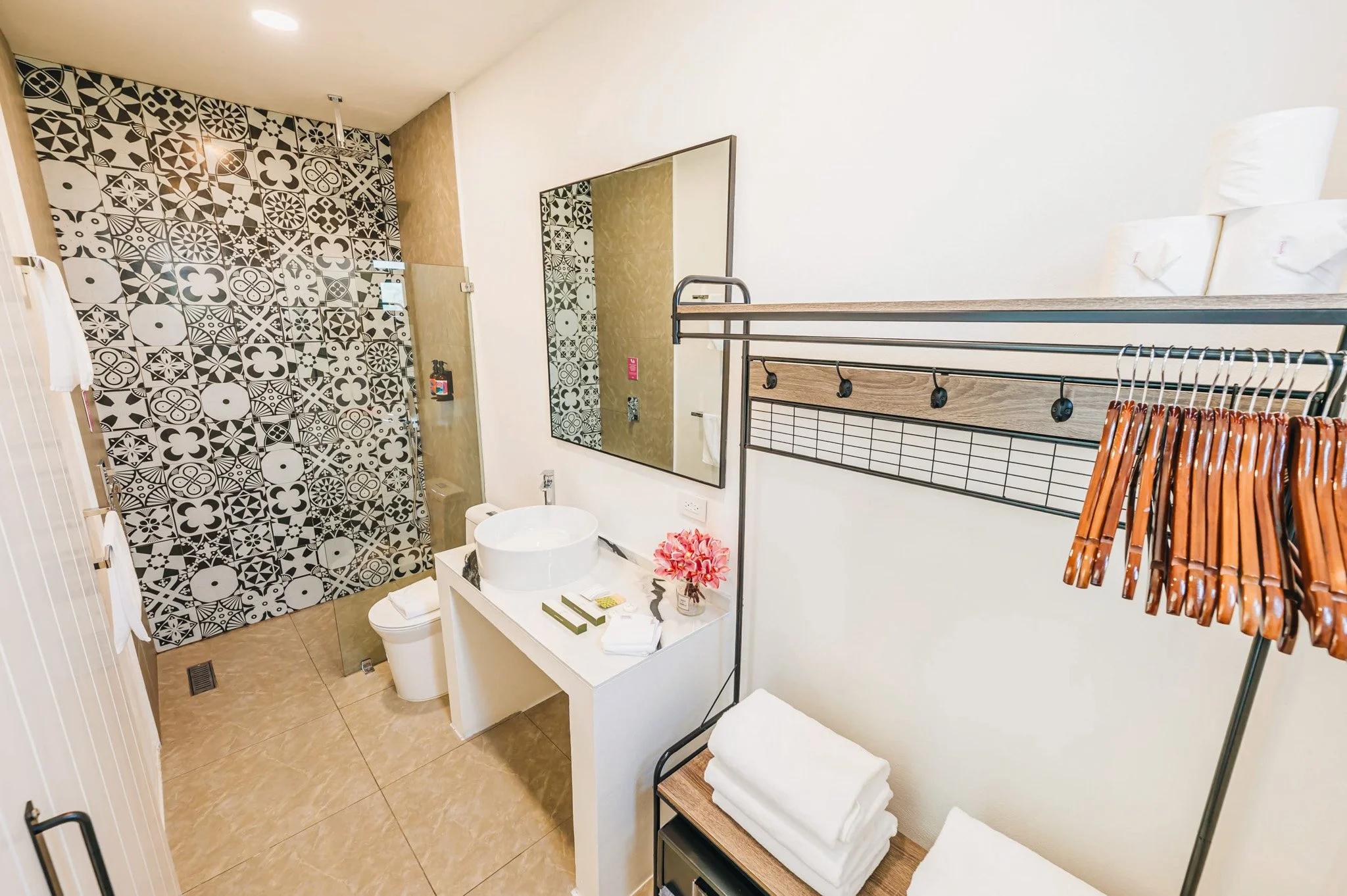 Modern bathroom with patterned black and white shower wall, white toilet, round vessel sink on a white vanity, mirror, and towel rack with hangers and towels.