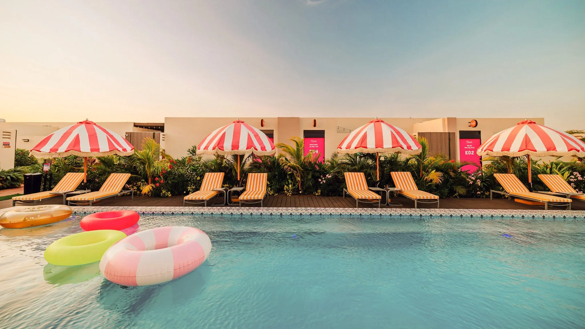 Poolside scene with six orange and white striped lounge chairs under six red and white striped umbrellas, surrounded by tropical plants, with floating inflatable rings in the pool in the foreground, and a building with pink signs in the background du