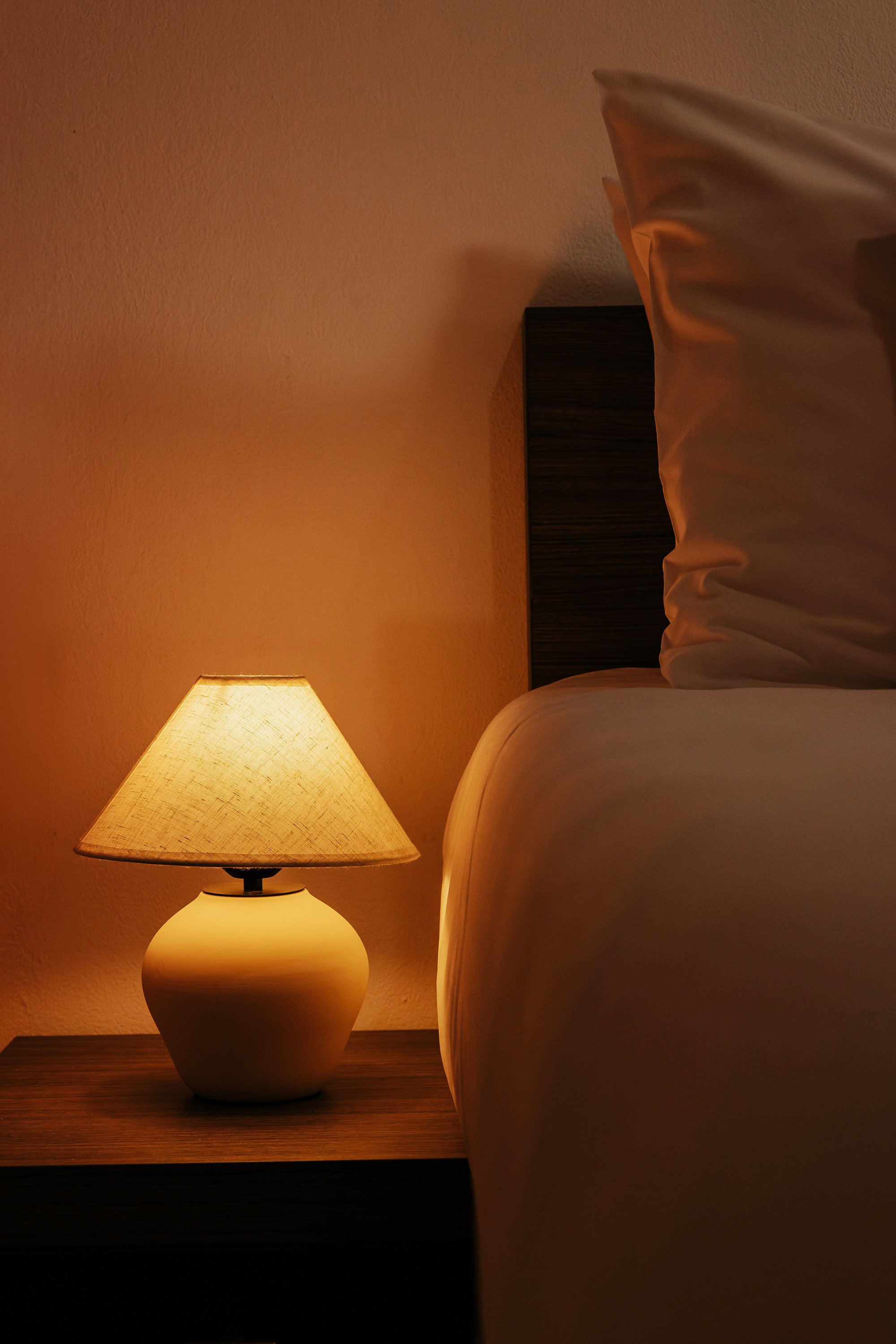 A bedside lamp on a wooden nightstand next to a bed with white sheets and pillows in a softly lit room.