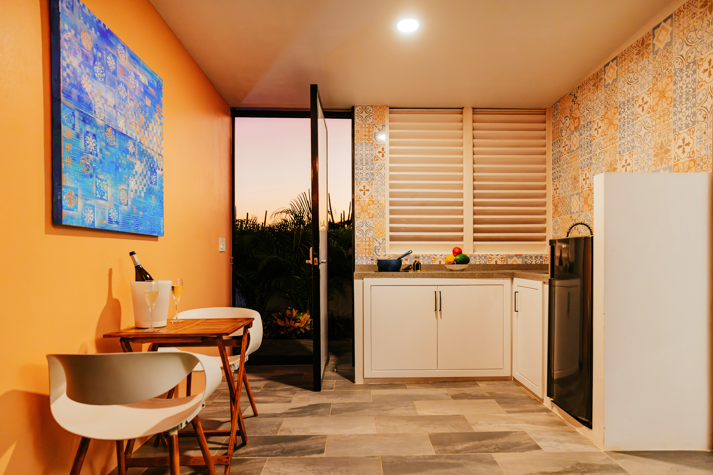 Cozy kitchen with orange wall, blue artwork, white cabinets, and patterned tile accents; open door leading to outdoor sunset.