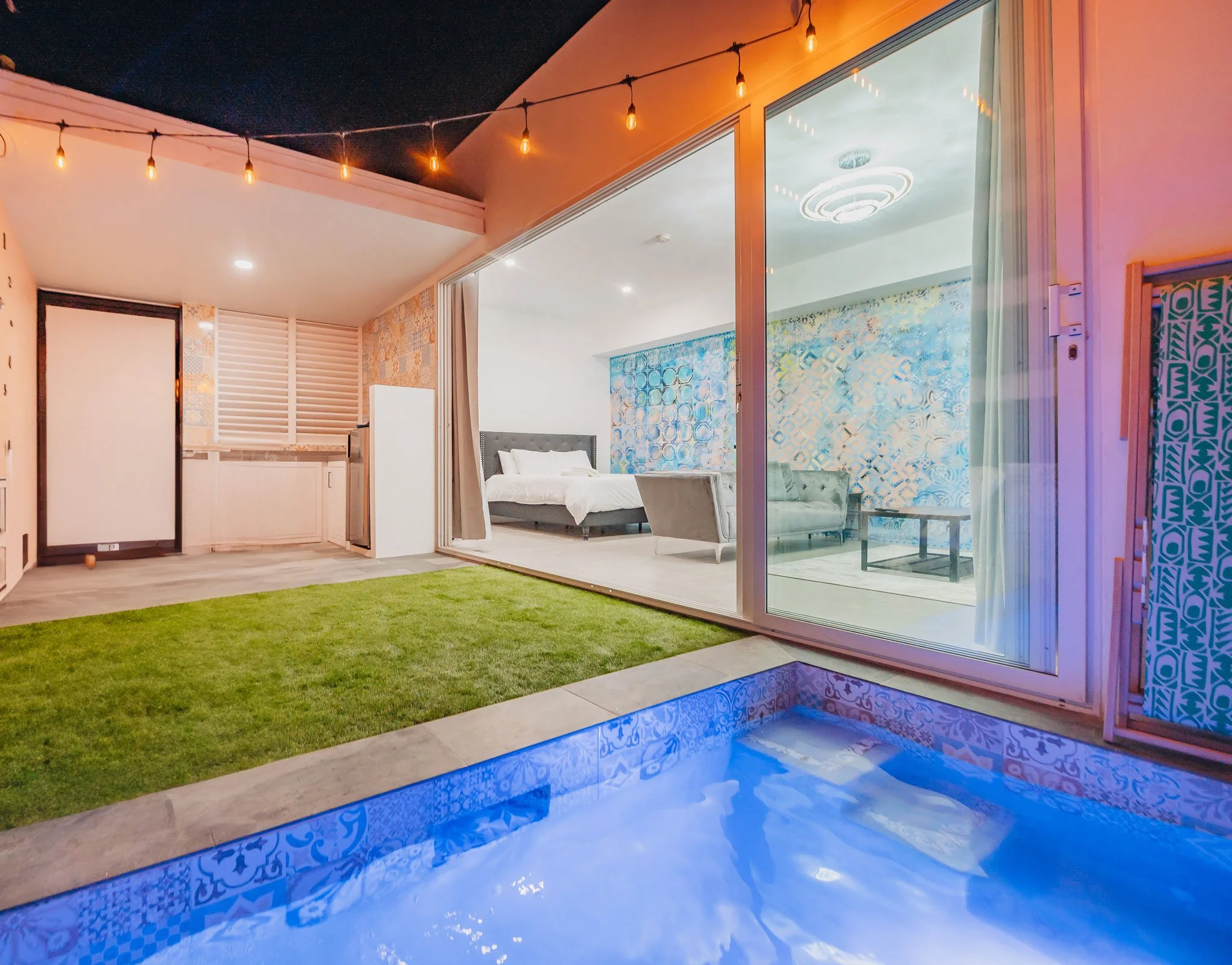 View of a modern indoor-outdoor living space with a small pool, glass sliding door, bedroom and living room, and decorative lighting.