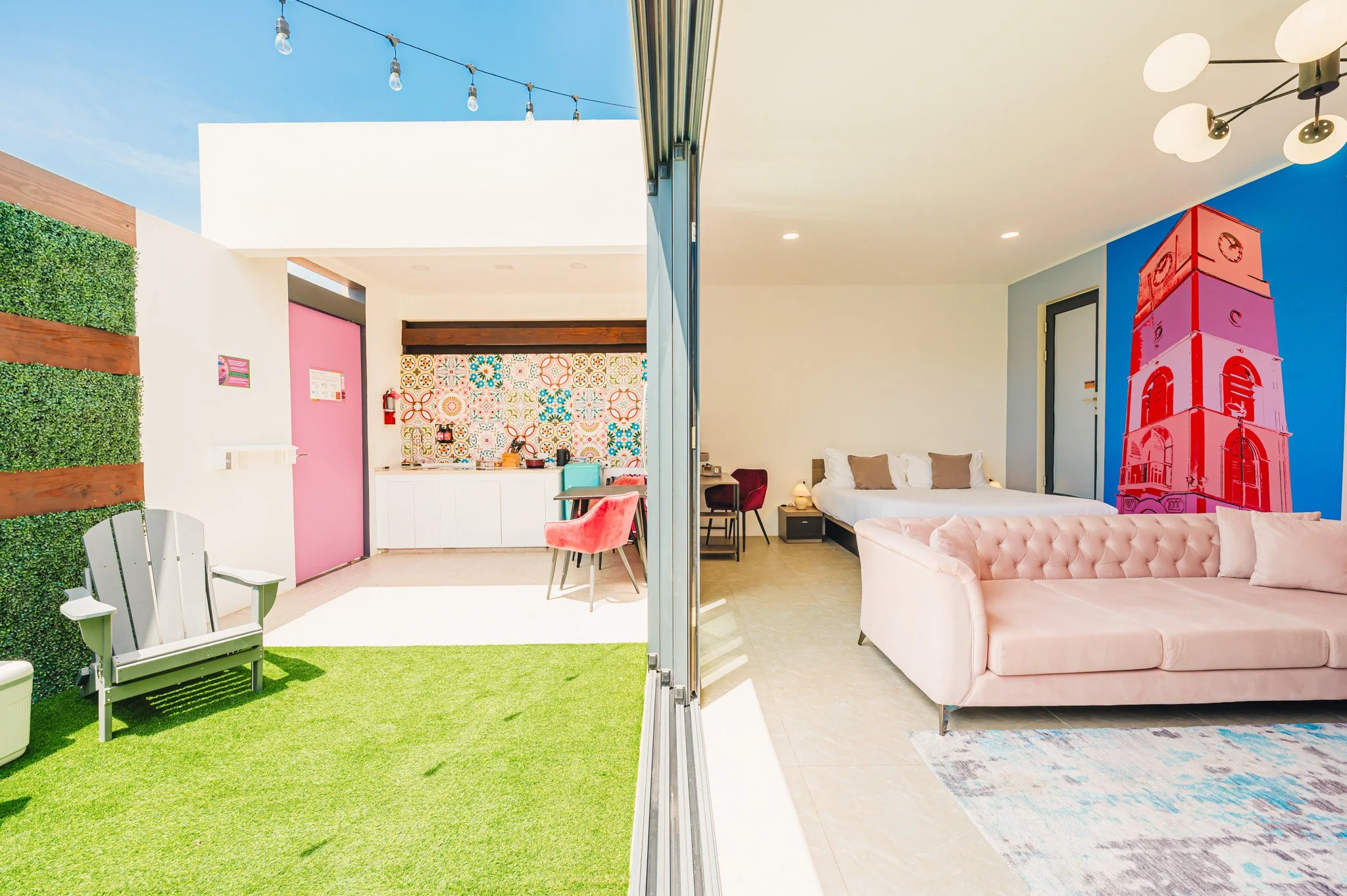 Split view of an indoor and outdoor space of a modern apartment, outdoor features a small green lawn, patio furniture, and string lights; indoor features a bright bedroom and colorful wall art, with a tall pink tower painting on the wall.
