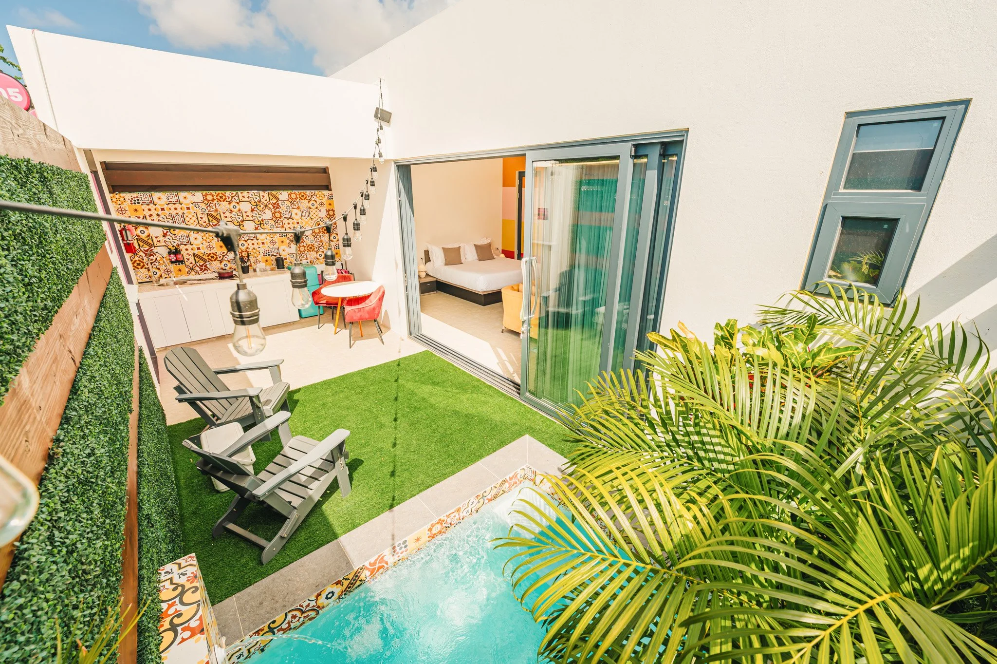 Outdoor patio area with a small pool, green artificial grass, wooden Adirondack chairs, and a string of outdoor lights. There is a sliding glass door leading to an indoor bedroom with a bed and sitting area. Tropical plants are also visible.