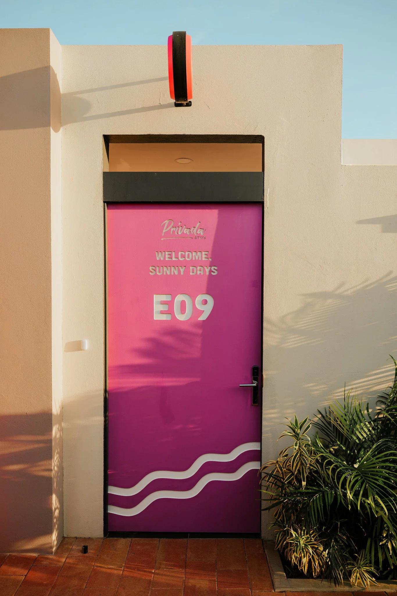 Pink door with white text that says "Privada Stay's Welcome Sunny Days E09," with decorative white wavy lines at the bottom, surrounded by a beige wall and green plants on the right. A black and red light fixture is mounted above the door.