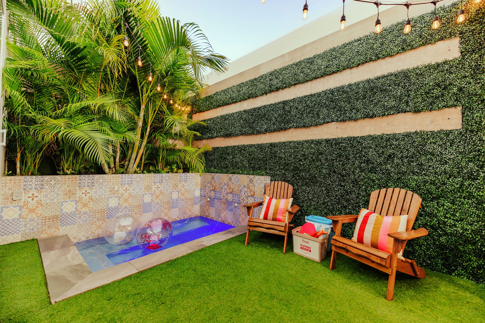 Small backyard with green grass, two wooden chairs with striped cushions, a small decorative pool with two floating balls, lush green plants, a decorative tiled wall, and string lights hanging above, in a modern outdoor setting.