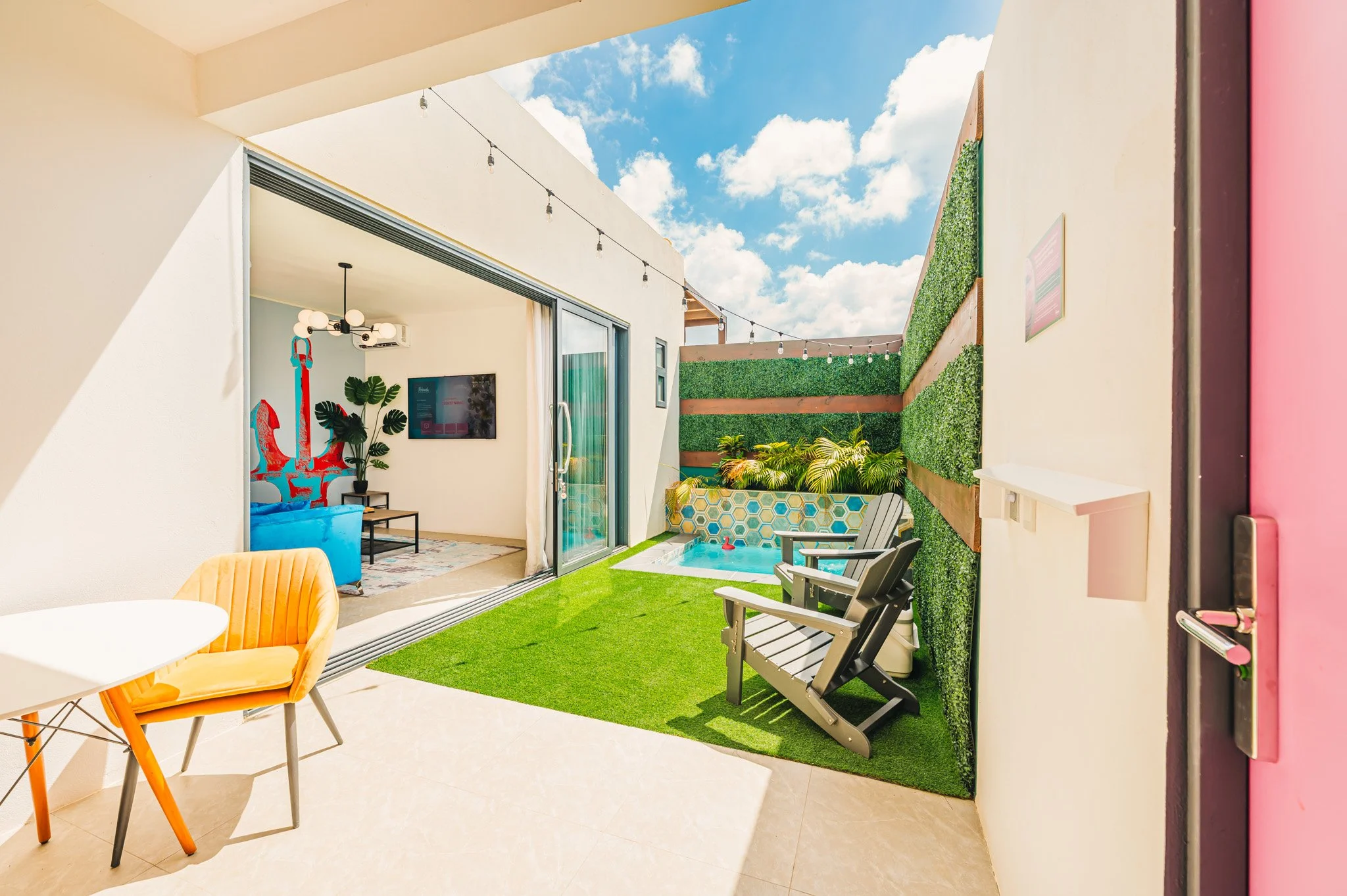 Indoor-outdoor living space with a patio, artificial grass, small pool, lounge chairs, a small table, and green plant wall. Inside, there is a room with a blue sofa, a wall-mounted TV, and decorative plants.