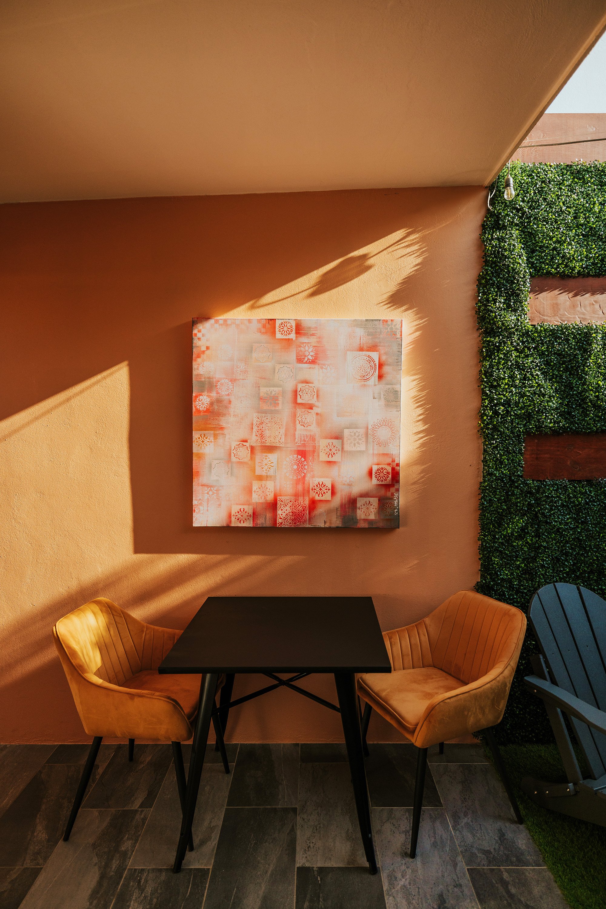 A cozy outdoor seating area with two mustard yellow velvet chairs, a black table, and an abstract red and white painting on a peach-colored wall. There is a green hedge on the right and sunlight casting shadows.