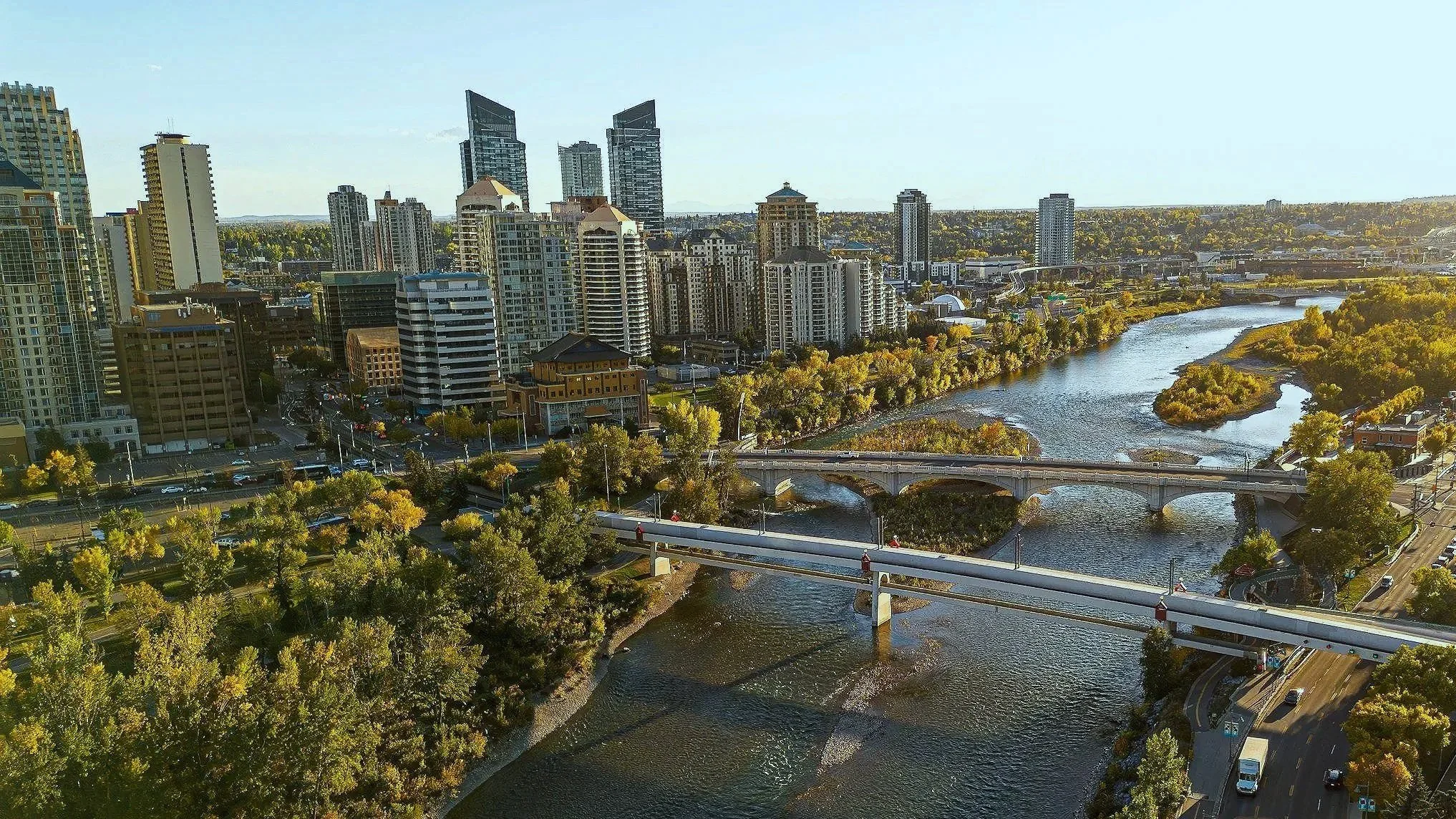 A city skyline with tall modern buildings next to a winding river and two bridges, with green trees and roads in the foreground.