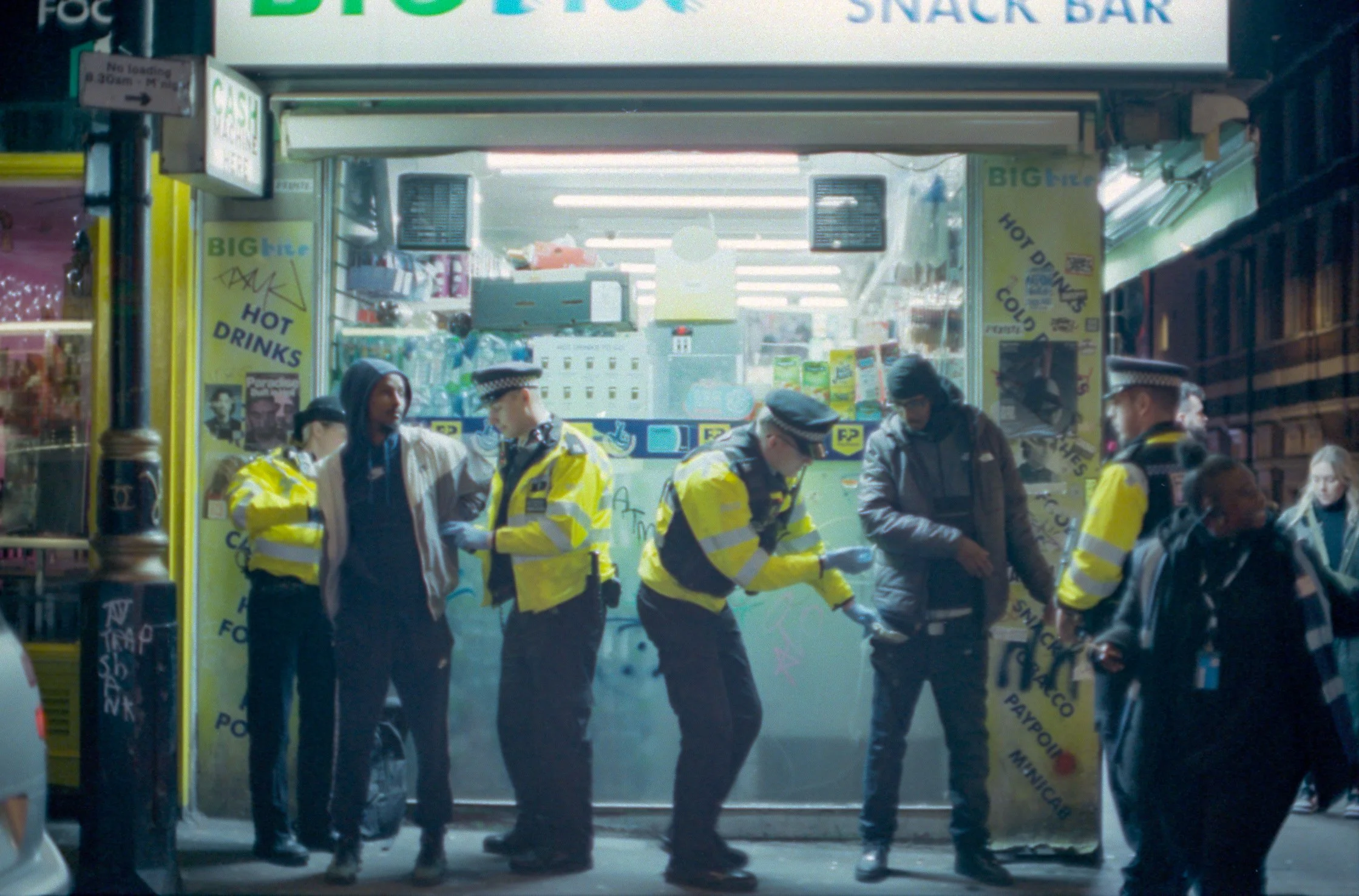 Group of police officers and civilians standing outside a small corner store on a city street at night. The store has signs for snacks, hot and cold drinks, and a cash register visible inside.