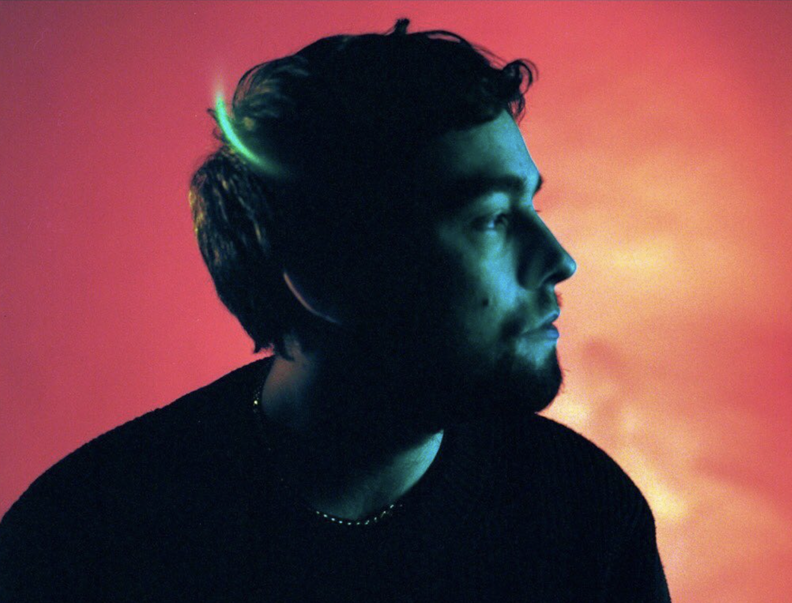 Side profile of a man with a beard and medium-length hair, illuminated with colorful lighting, against a pink and orange background.