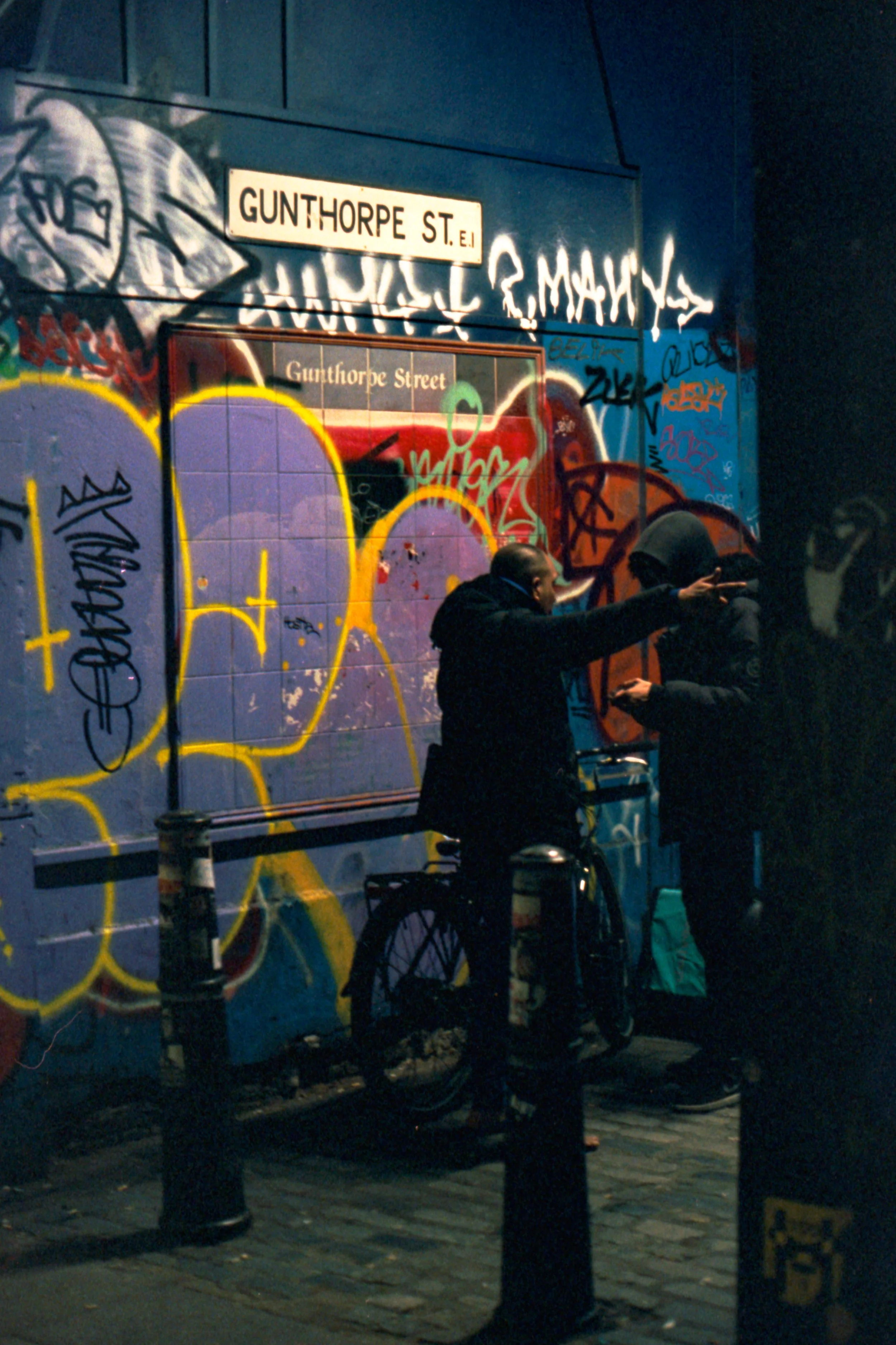 Two men having a conversation on a graffiti-covered street corner at night, with street signs reading 'GUNTHORPE ST. E.' and 'GUNTHORPE Street' visible, and a bicycle leaning against the wall.