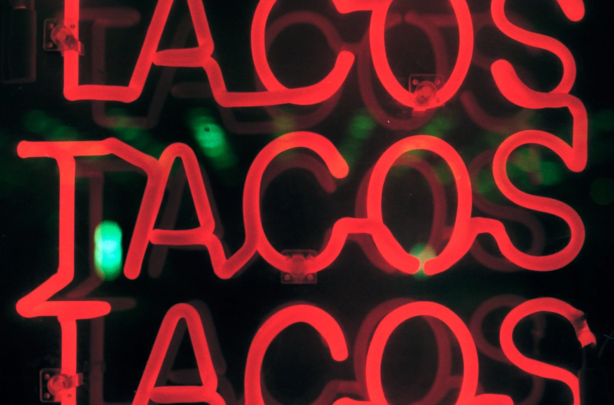 Red neon sign that reads 'TACOS' reflected in a mirror, with some green bokeh lights in the background.