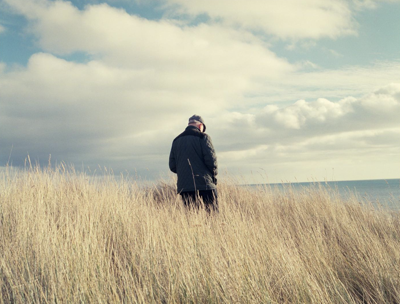 A person standing alone in tall, golden grass near the ocean under a partly cloudy sky.