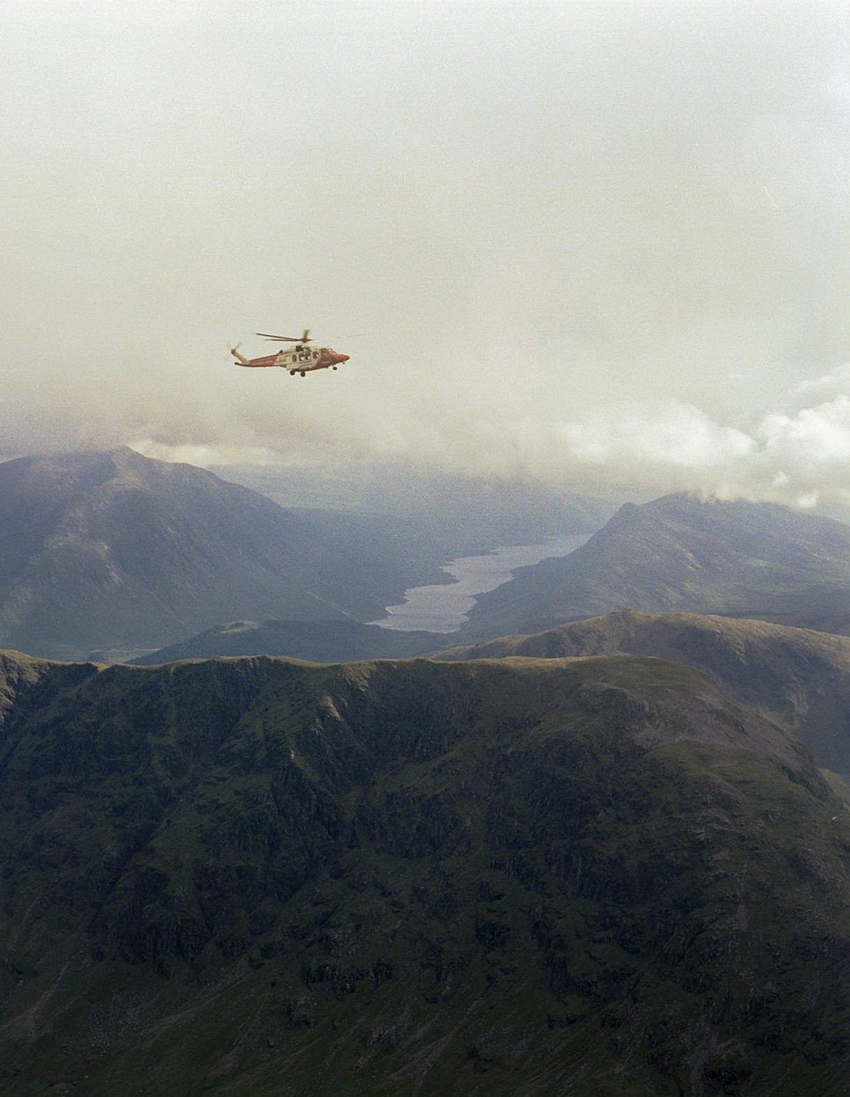 A red and white helicopter flying over green mountainous landscape with a lake in the distance under cloudy sky.