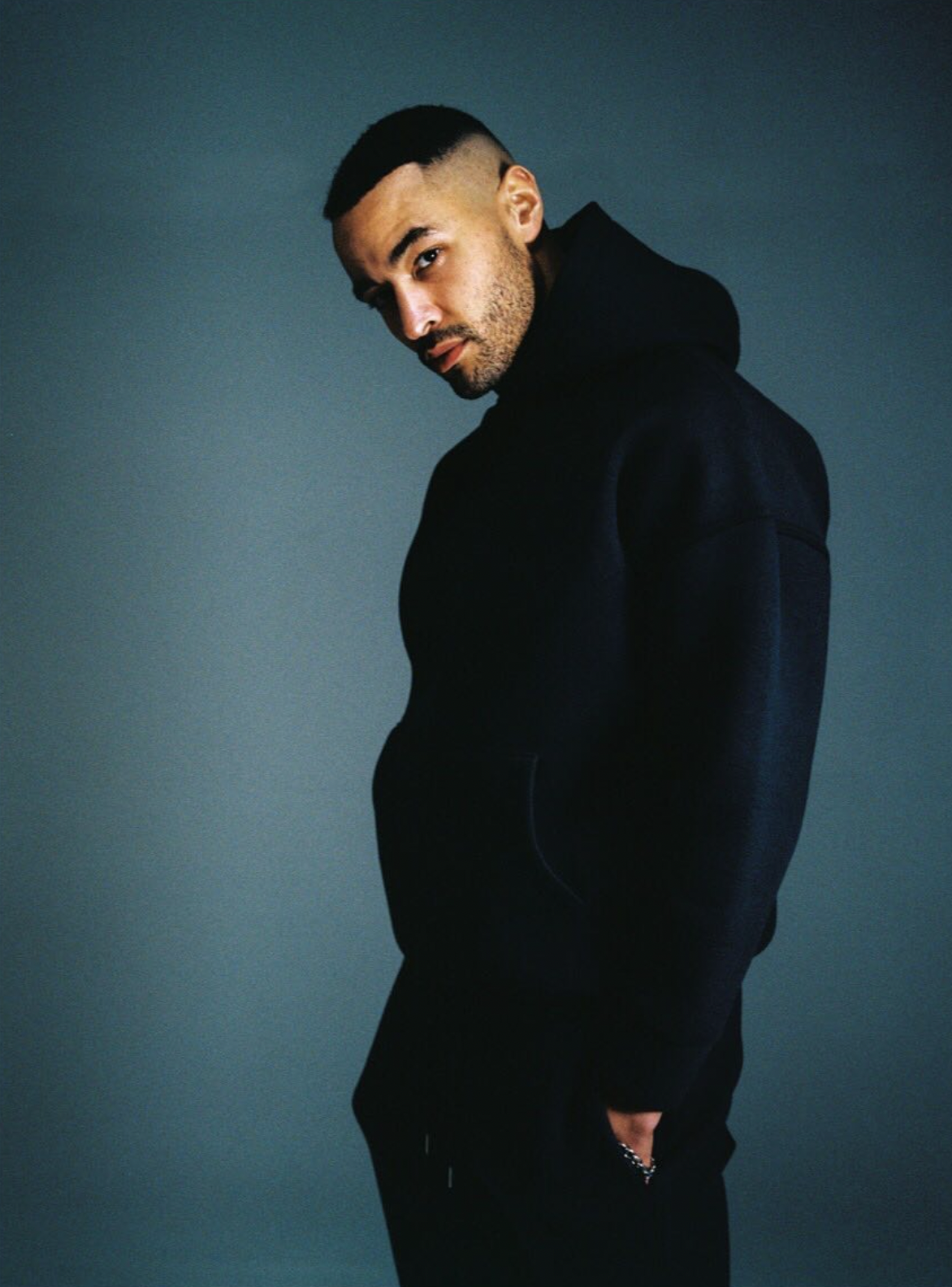 A man with a fade haircut, beard, wearing a black hoodie and black pants, stands against a plain, dark background, looking at the camera with a serious expression.