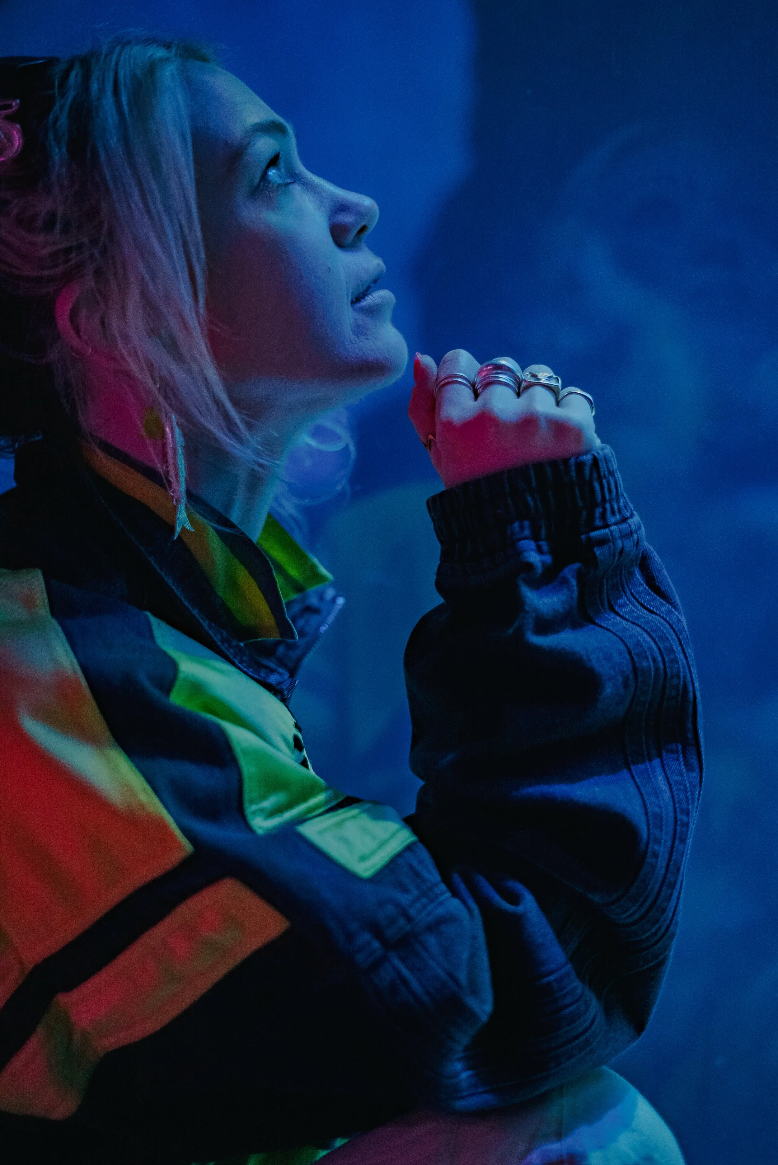 A woman with blonde hair is looking upward with a thoughtful expression, illuminated by blue and pink neon lights, wearing a dark high-collared jacket with a rainbow-colored pattern.
