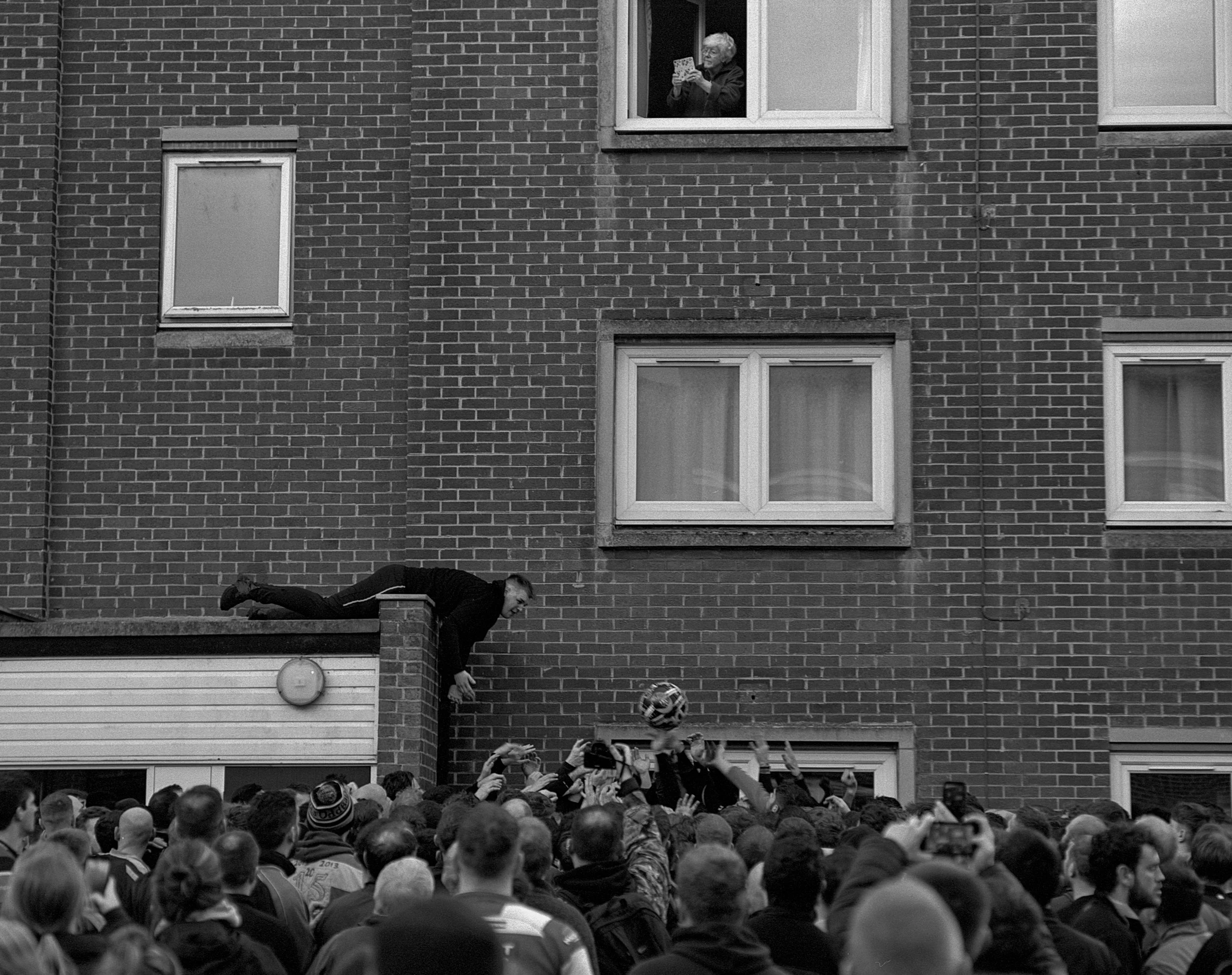 Crowd gathered outside a brick building with windows, with a man leaning from a balcony reaching toward people below, some taking photos or videos.