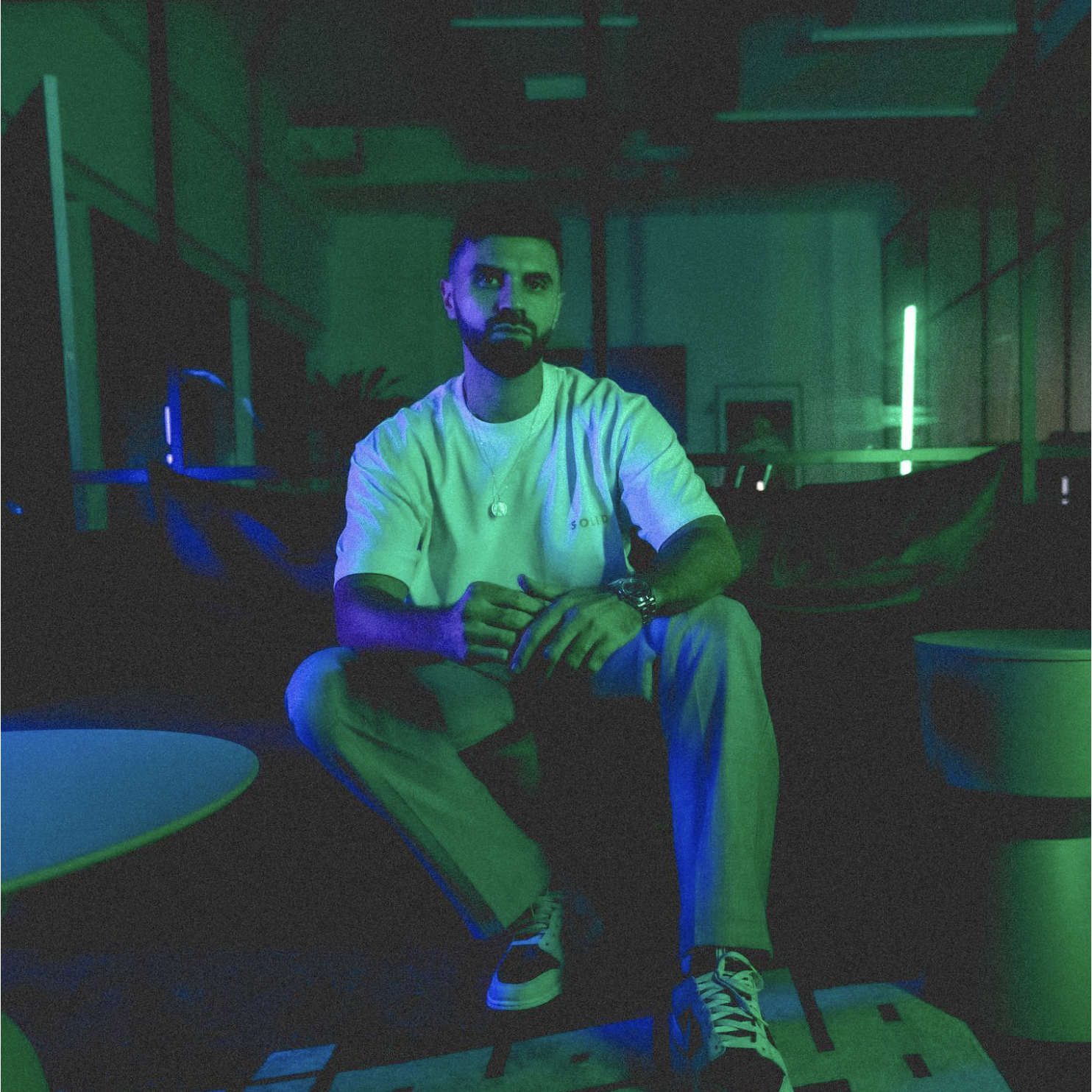 A man sitting in a dimly lit room with green and blue lighting, wearing a white T-shirt and light-colored pants.