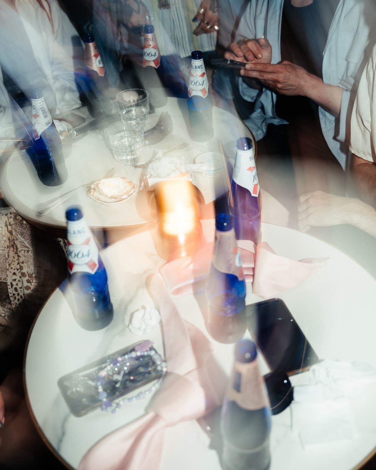 Reflection of a social gathering around a table with bottles, glasses, and a person using a smartphone, captured through a reflective surface.