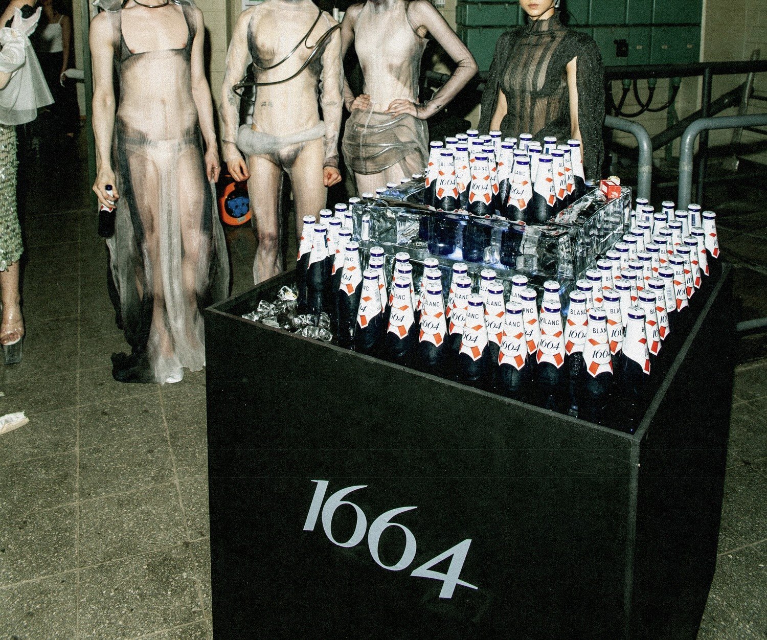 A display of bottles with bottle-shaped labels, arranged on a black table with a large '1604' logo on the side, surrounded by mannequins dressed in sheer, revealing clothing.