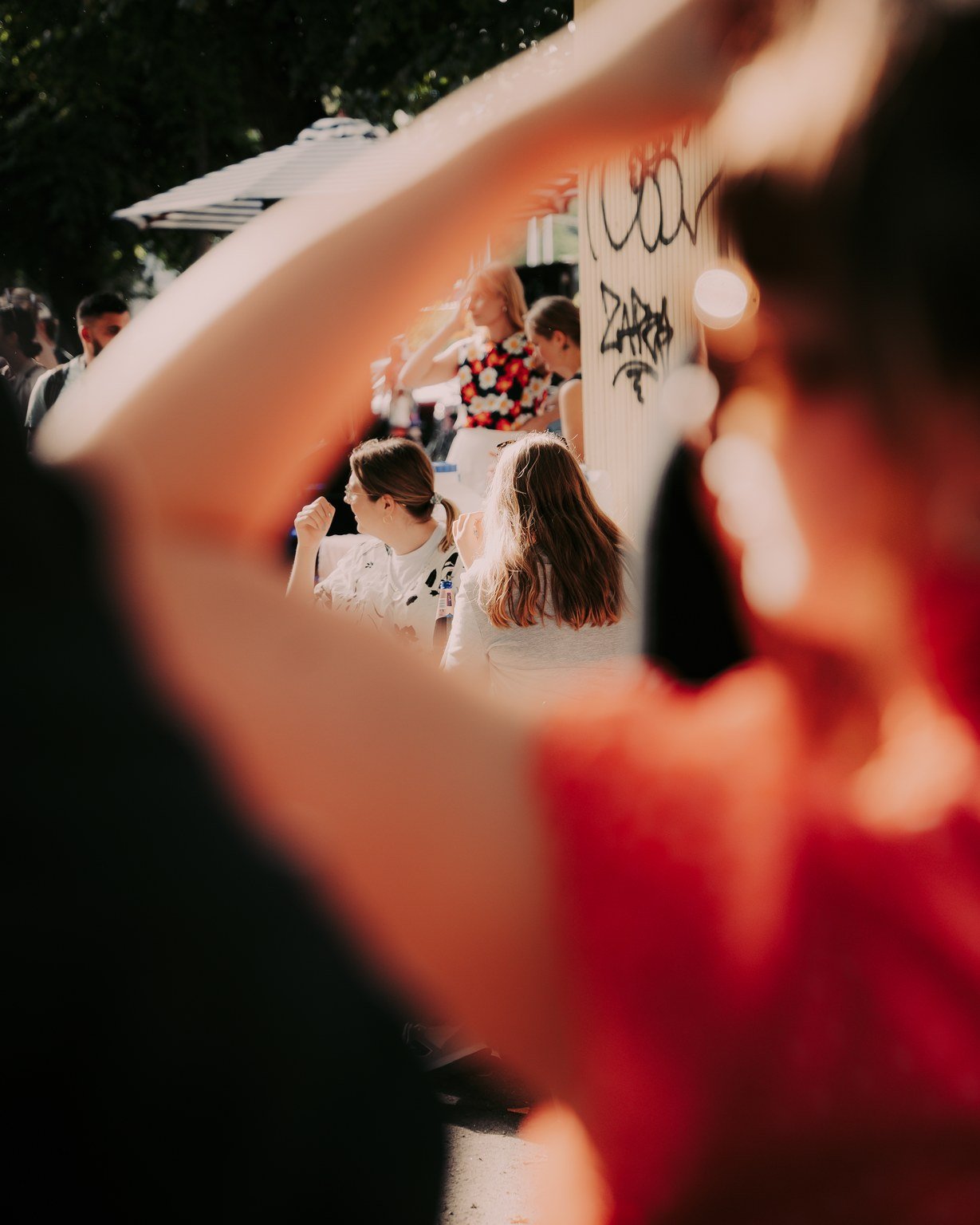 People sitting outdoors at a sunny event, with a view through a blurry, circular frame in the foreground, featuring a woman in a floral dress and others engaged in conversation.