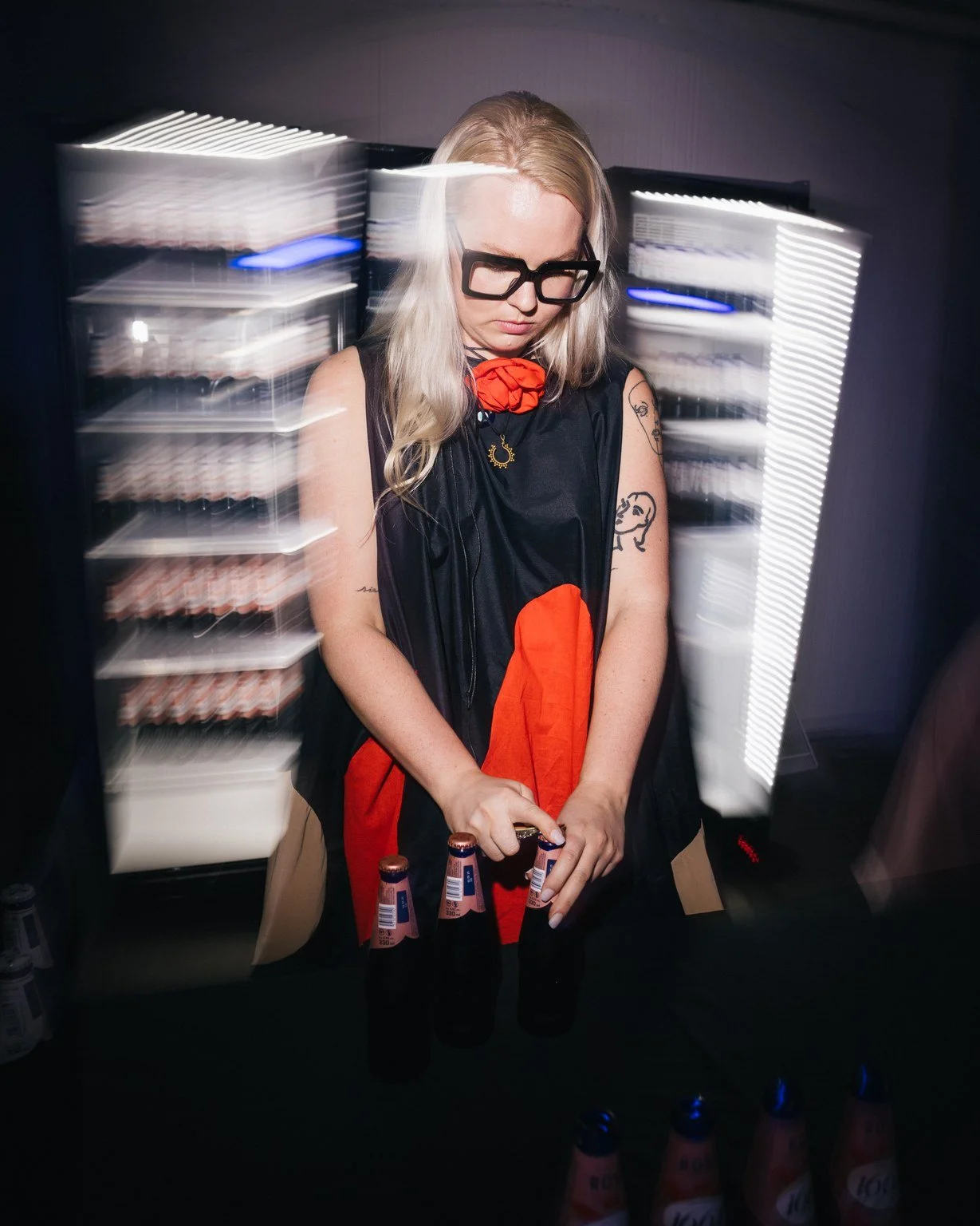 A woman with blonde hair, wearing glasses and a black and red dress, standing behind a table with beer bottles, in a room with light streaks due to motion blur.