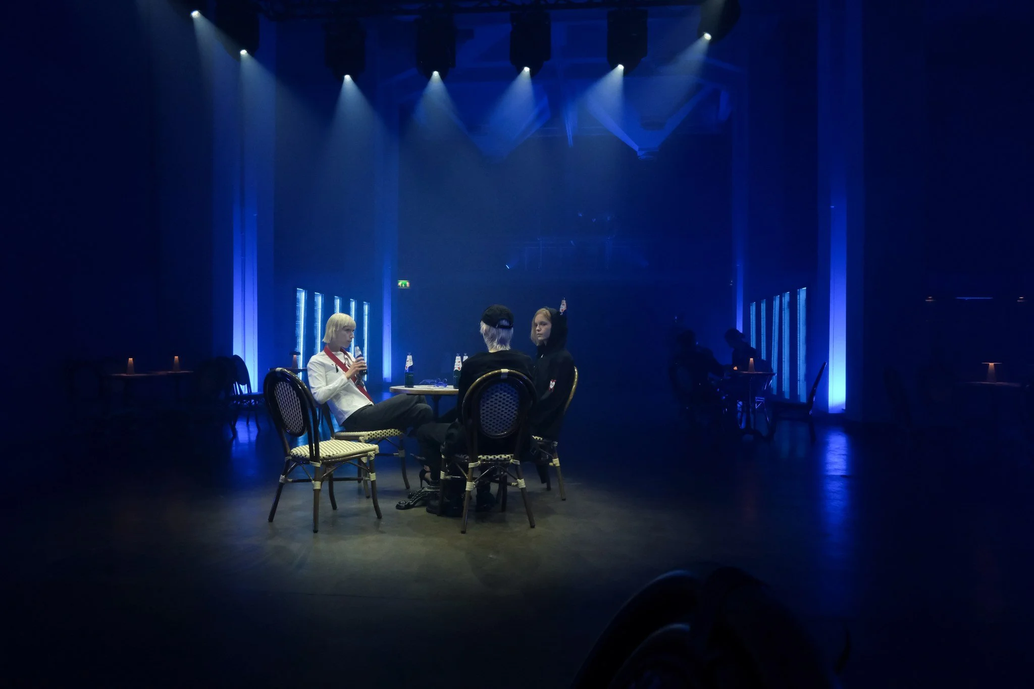 Three people sitting at a table in a dimly lit, blue-lit room, with two bottles of beer on the table, others seated at tables in the background.