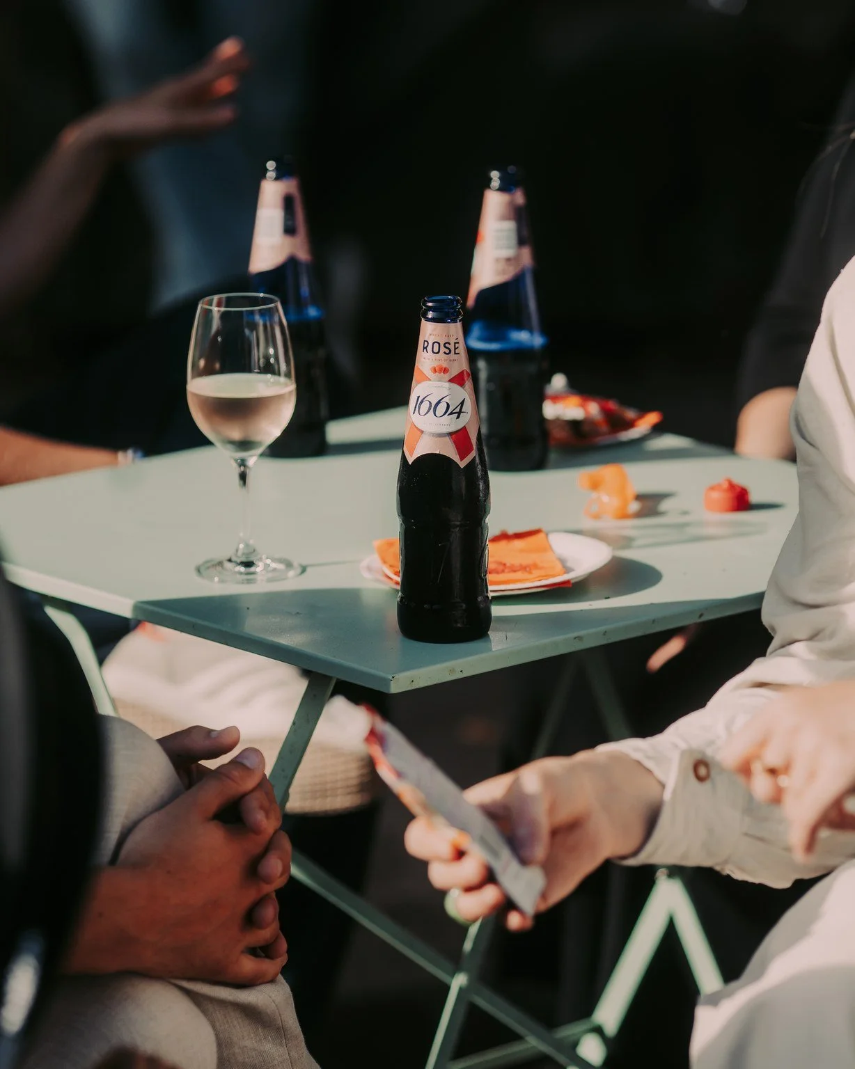 People sitting around a table with bottles of rosé beer, a wine glass, and snacks while one person uses a smartphone.