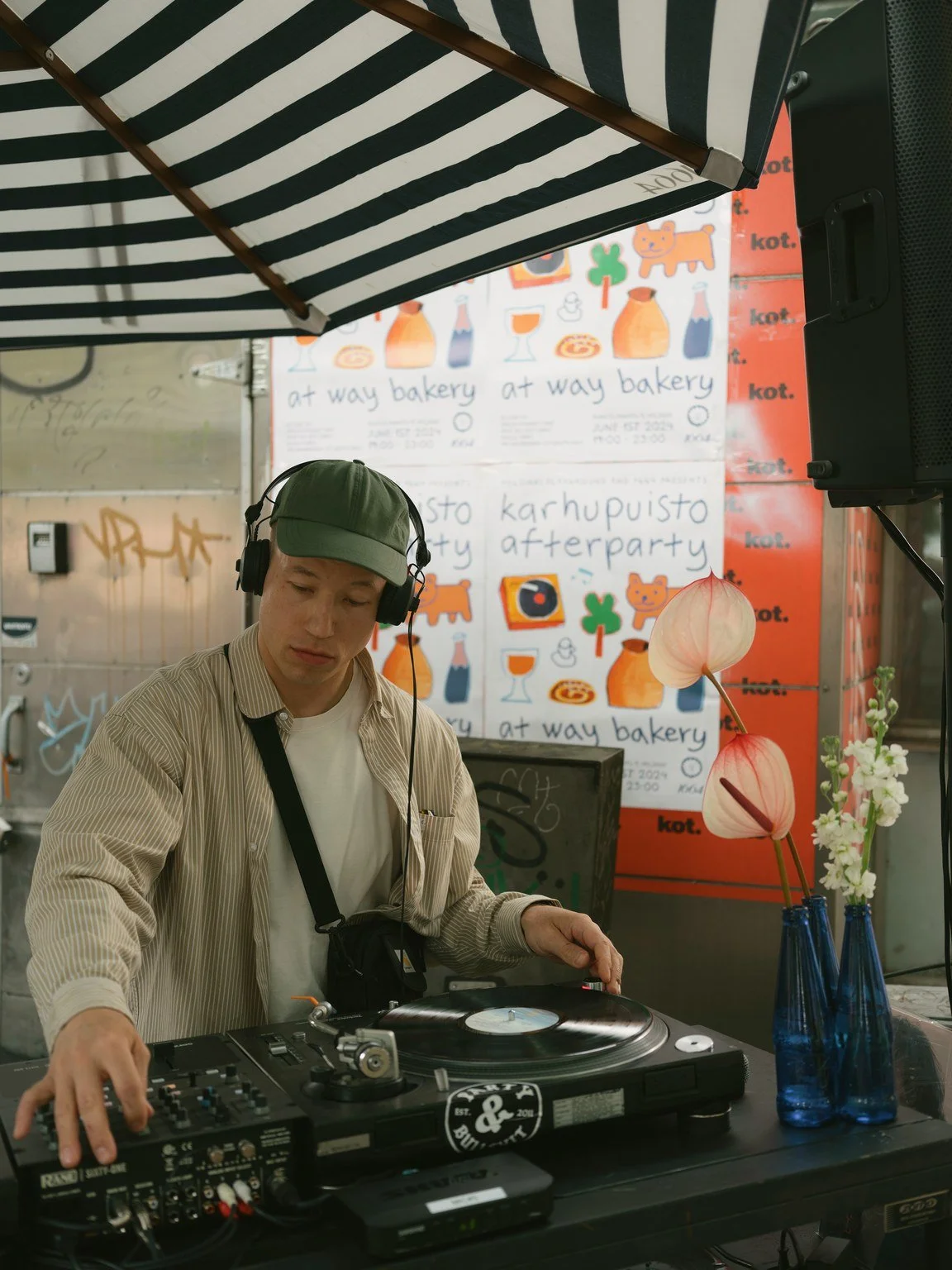 A young man wearing a green cap and headphones is DJing with a turntable and mixer at an outdoor event. Behind him, there is a colorful sign with drawings of bottles, jars, and animals, and handwritten text advertising a bakery and afterparty.