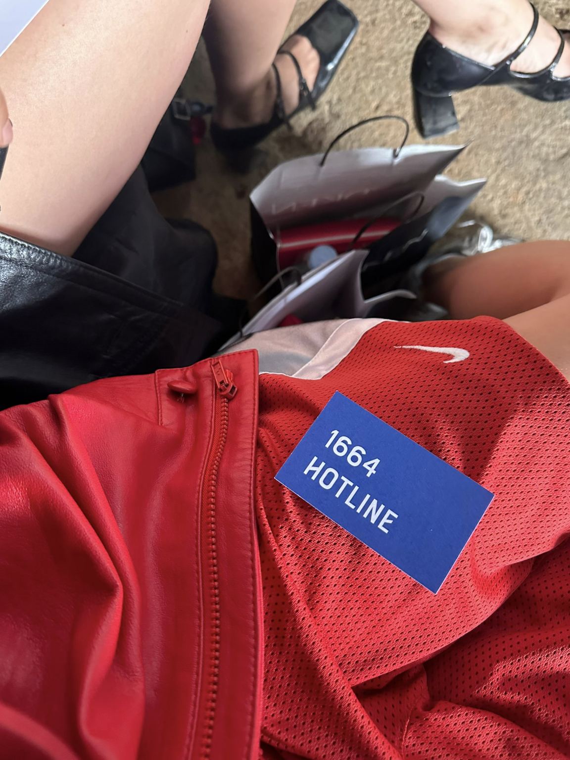 Person wearing a red sports jacket with a blue tag reading '1664 HOTLINE' sitting on a seat, with two other people nearby. Shoes and shopping bags are visible on the ground.