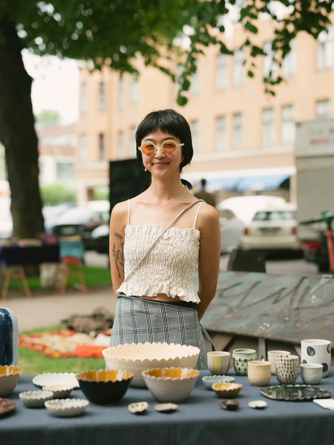 Woman selling handmade pottery at an outdoor market, standing behind a table filled with ceramic bowls and cups, smiling with sunglasses, in a park with trees and buildings in the background.