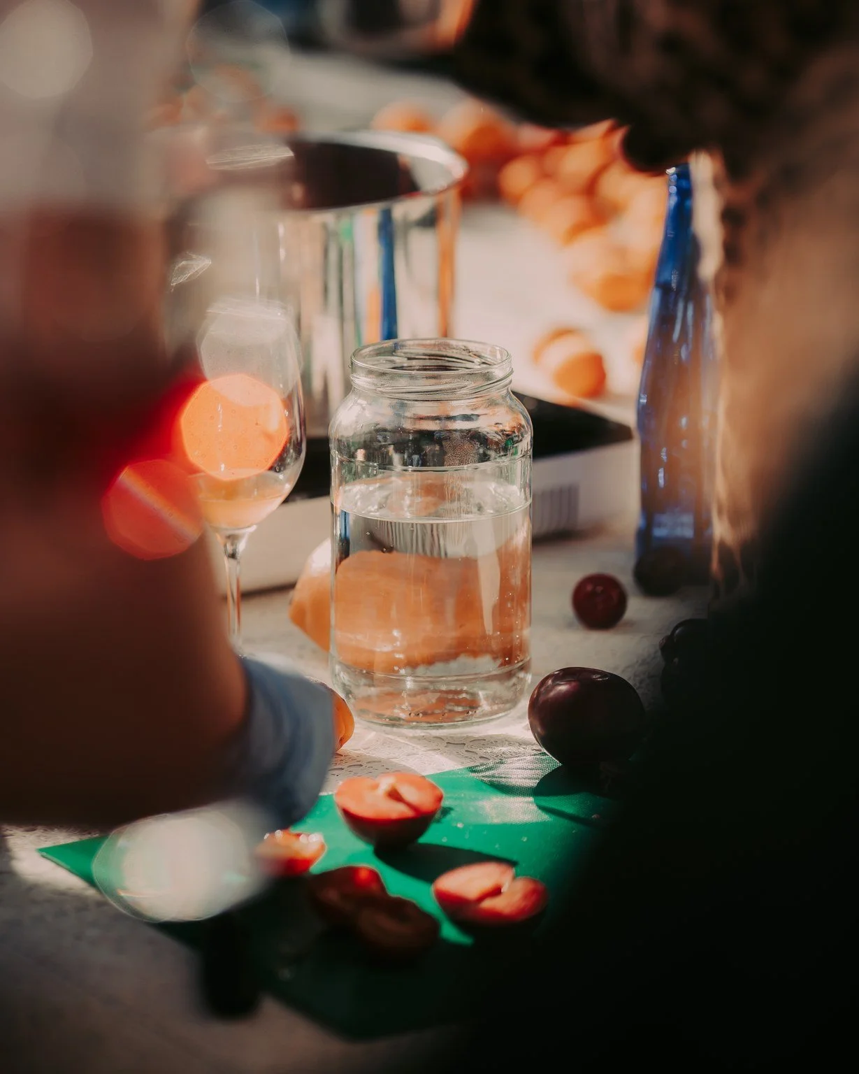 A close-up of a kitchen or dining table with sliced cherries on a green cutting board, a glass jar of water, a partially visible wine glass, a blue bag of chips, and some orange slices. Sunlight creates a warm, soft glow with bokeh effects in the bac
