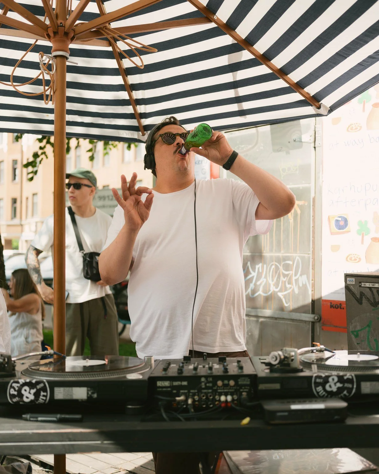 A DJ wearing headphones and sunglasses is drinking from a green bottle while standing behind a DJ setup at an outdoor venue, with a striped umbrella overhead and a man with tattoos and a cap in the background.